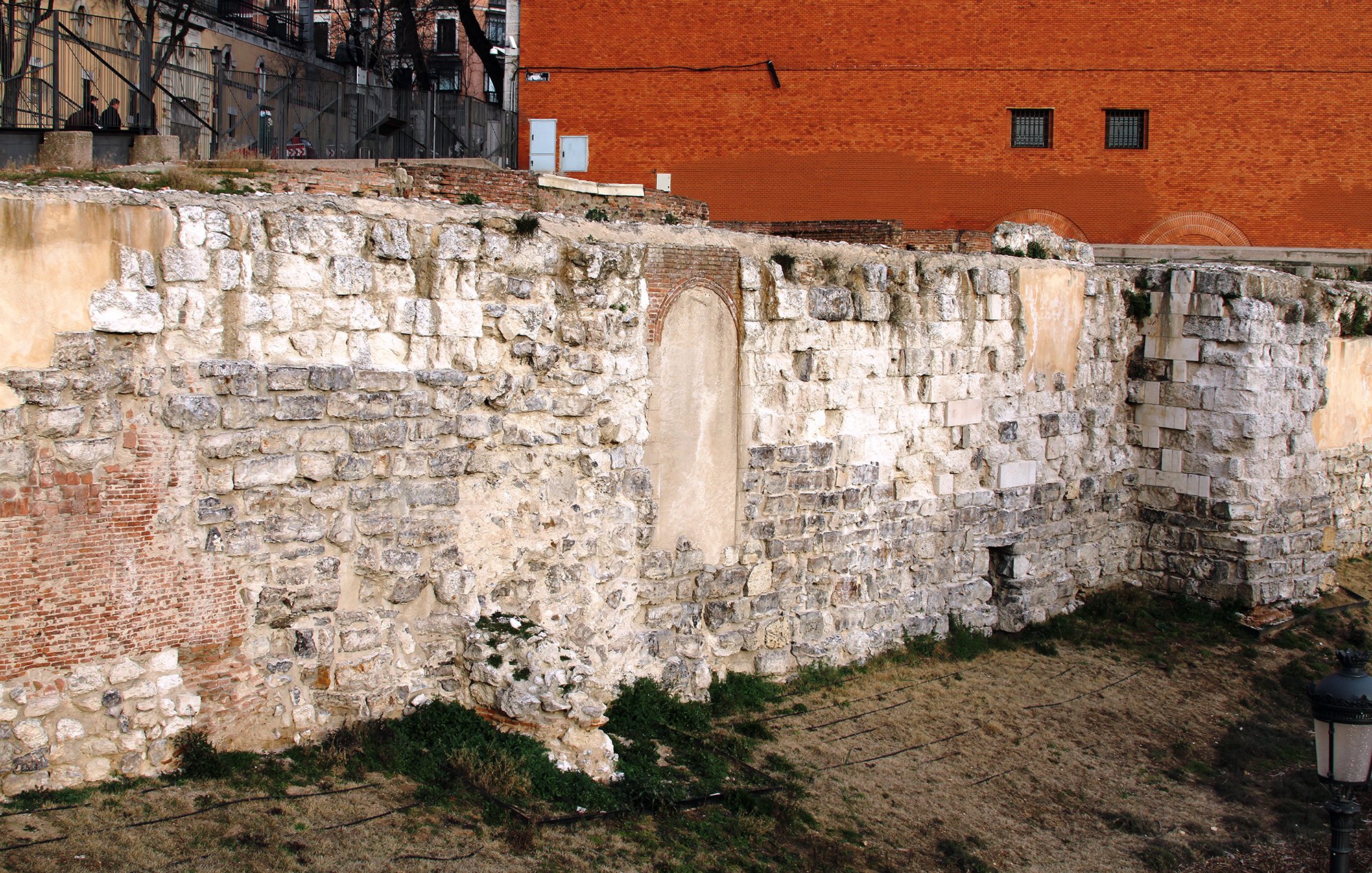L'image montre une section de mur en pierre, probablement une ancienne structure ou fortification. Les pierres sont de tailles et de formes variées, avec des parties couvertes de végétation. En arrière-plan, on peut apercevoir un mur de couleur rouge et quelques éléments de structure moderne. Le site semble être à l'extérieur, probablement dans une zone historique, avec une ambiance de délabrement et d'abandon.