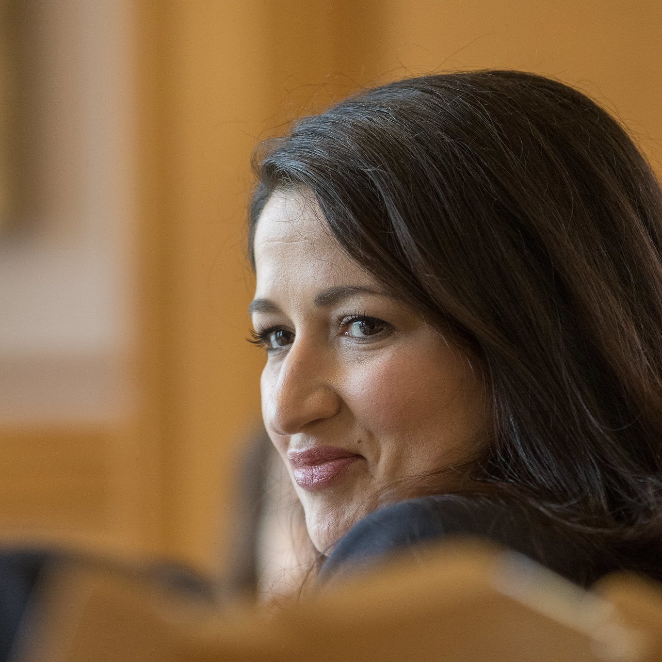 Je ne peux pas identifier les personnes dans l'image. Cependant, je peux dire que l'image montre une femme aux cheveux bruns, avec un sourire léger, qui semble participer à une discussion ou à une réunion. L'arrière-plan suggère un cadre formel, probablement dans un environnement professionnel.