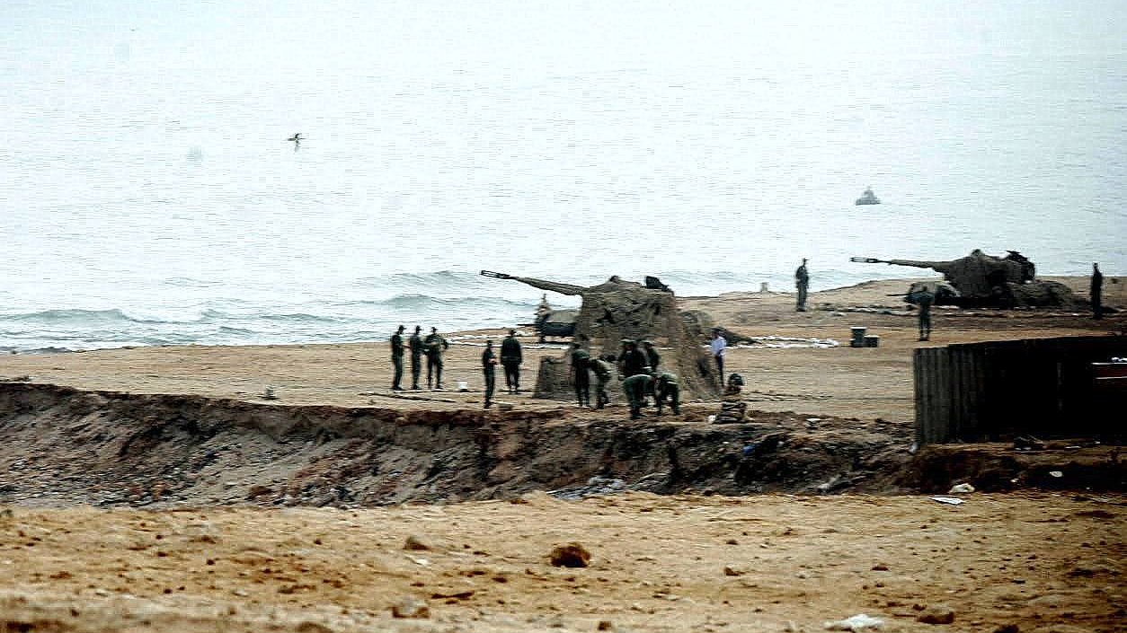 L'image montre une scène côtière où des militaires sont visibles sur une plage. On aperçoit des canons posés sur le sable, ainsi qu'un groupe de soldats qui semblent s'affairer autour. Le paysage est brumeux, avec une mer calme au fond et un ciel nuageux. L'atmosphère paraît sérieuse et militaire, avec une concentration sur les préparatifs ou les opérations en cours.