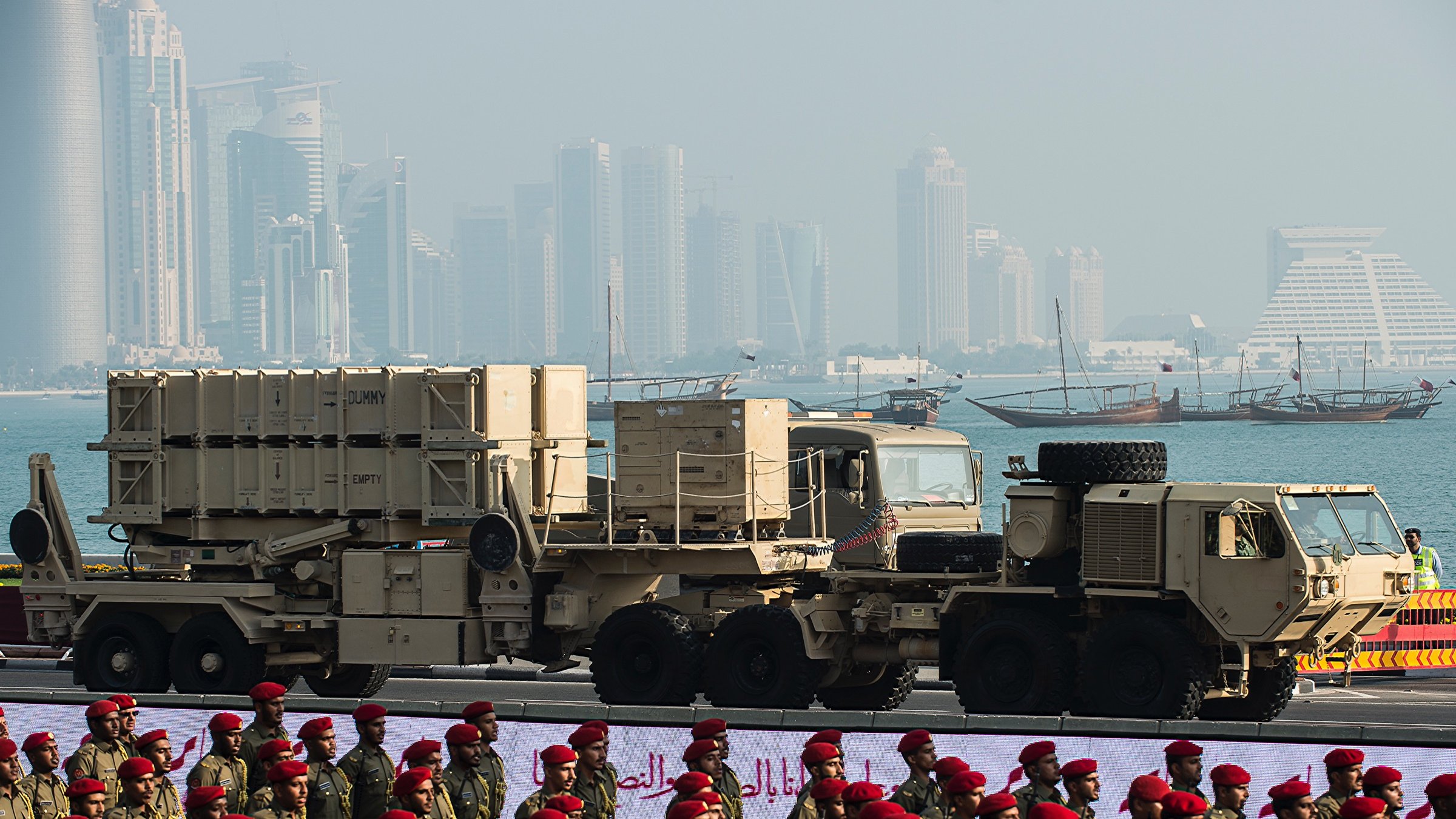 L'image montre des équipements militaires, probablement des systèmes de défense aérienne, positionnés sur des camions. En arrière-plan, on aperçoit une ville moderne avec des gratte-ciels et des bâtiments contemporains, ainsi qu'une étendue d'eau et des bateaux traditionnels. Au premier plan, des soldats en uniforme, peut-être d'une force armée, sont alignés, ajoutant une dimension cérémonielle à la scène. L'atmosphère semble être celle d'un défilé militaire ou d'un événement similaire.