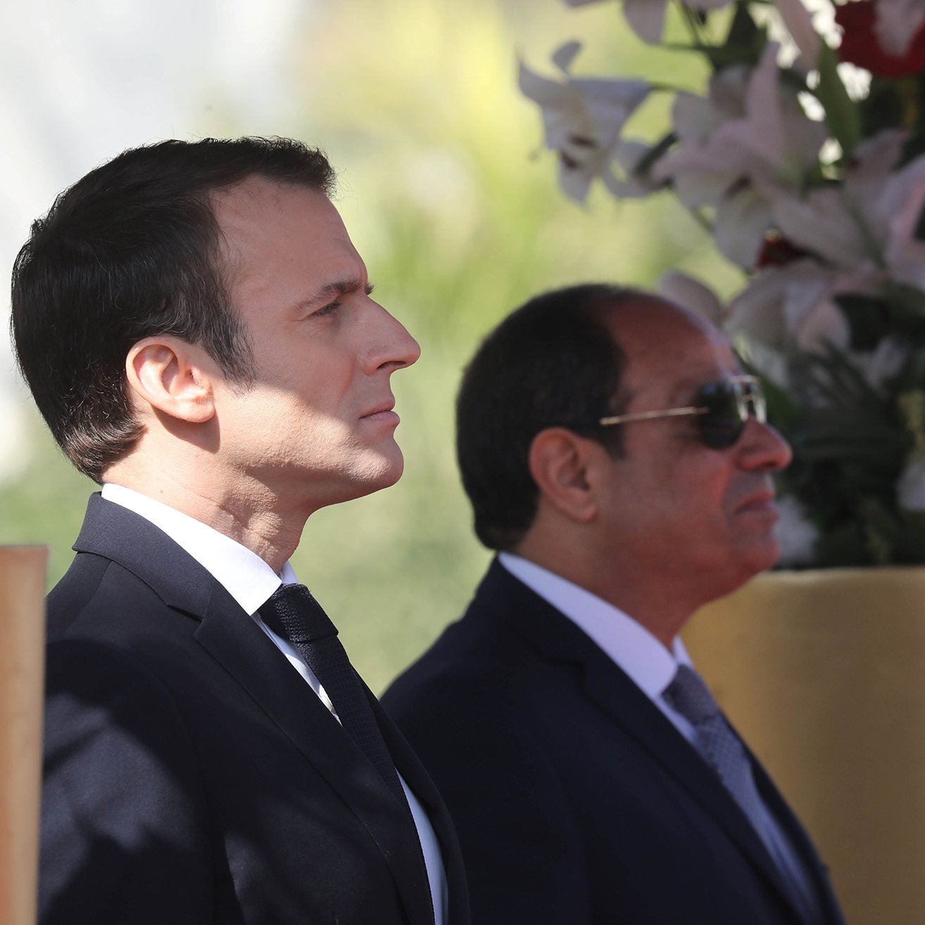 L'image montre deux hommes debout, apparemment lors d'un événement officiel. L'un d'eux est en costume sombre avec une cravate, il a une expression concentrée et regarde droit devant lui. L'autre homme, également en costume, porte des lunettes de soleil et semble observateur. Ils se tiennent près de décorations florales, suggérant un cadre cérémonieux.