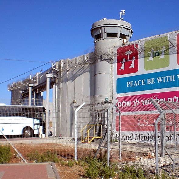 L'image montre un mur imposant, probablement un mur de séparation, avec une tour de surveillance au sommet. À la base du mur, il y a une grande pancarte qui affiche des messages liés à la paix, écrits en plusieurs langues. L'environnement est urbain, et on peut voir un bus stationné à proximité. Le tout est entouré d'une clôture barbelée, ce qui donne une impression de sécurité renforcée. Le ciel est dégagé et ensoleillé.