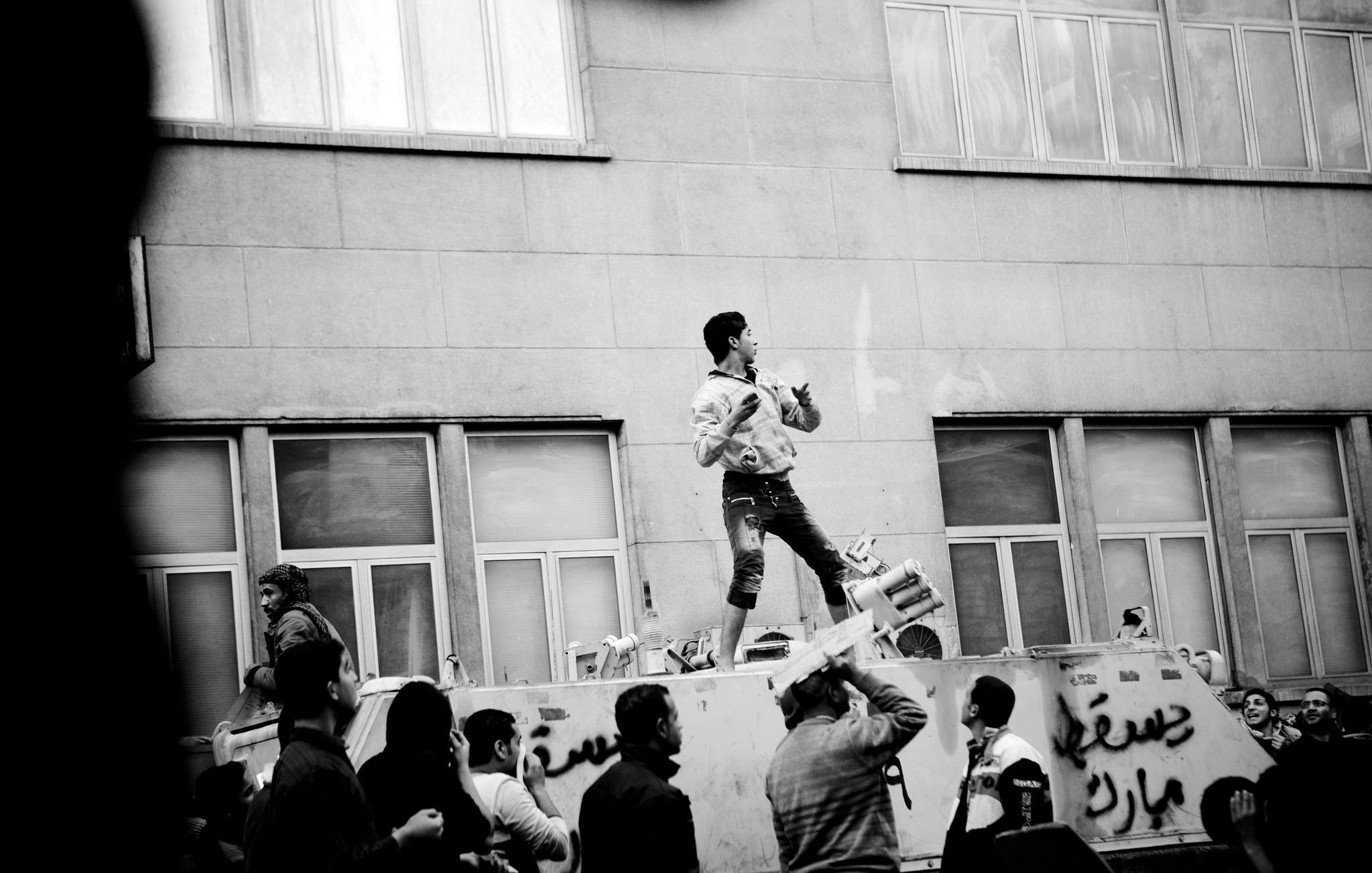 L'immagine mostra una scena di protesta in bianco e nero. Un giovane si trova in cima a un veicolo, apparentemente utilizzando un megafono o uno strumento, mentre sembra incoraggiare la folla sottostante. La gente intorno a lui è visibilmente coinvolta, alcuni alzano le mani o applaudono. Sul veicolo ci sono delle scritte, e l'architettura sullo sfondo suggerisce un contesto urbano. L'atmosfera è carica di energia e di determinazione.