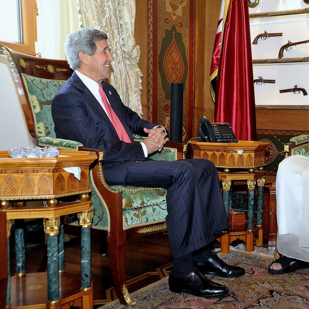 L'image montre deux hommes assis dans un bureau élégant, décoré avec des motifs riches et des couleurs vives. L'un des hommes porte un costume formel et sourit, tandis que l'autre est vêtu d'une tenue traditionnelle blanche, avec un chapeau noir. Ils semblent être en train de discuter dans une atmosphère amicale. En arrière-plan, on peut voir des objets décoratifs et des armes anciennes accrochées au mur, ajoutant à l'ambiance opulente de la pièce.