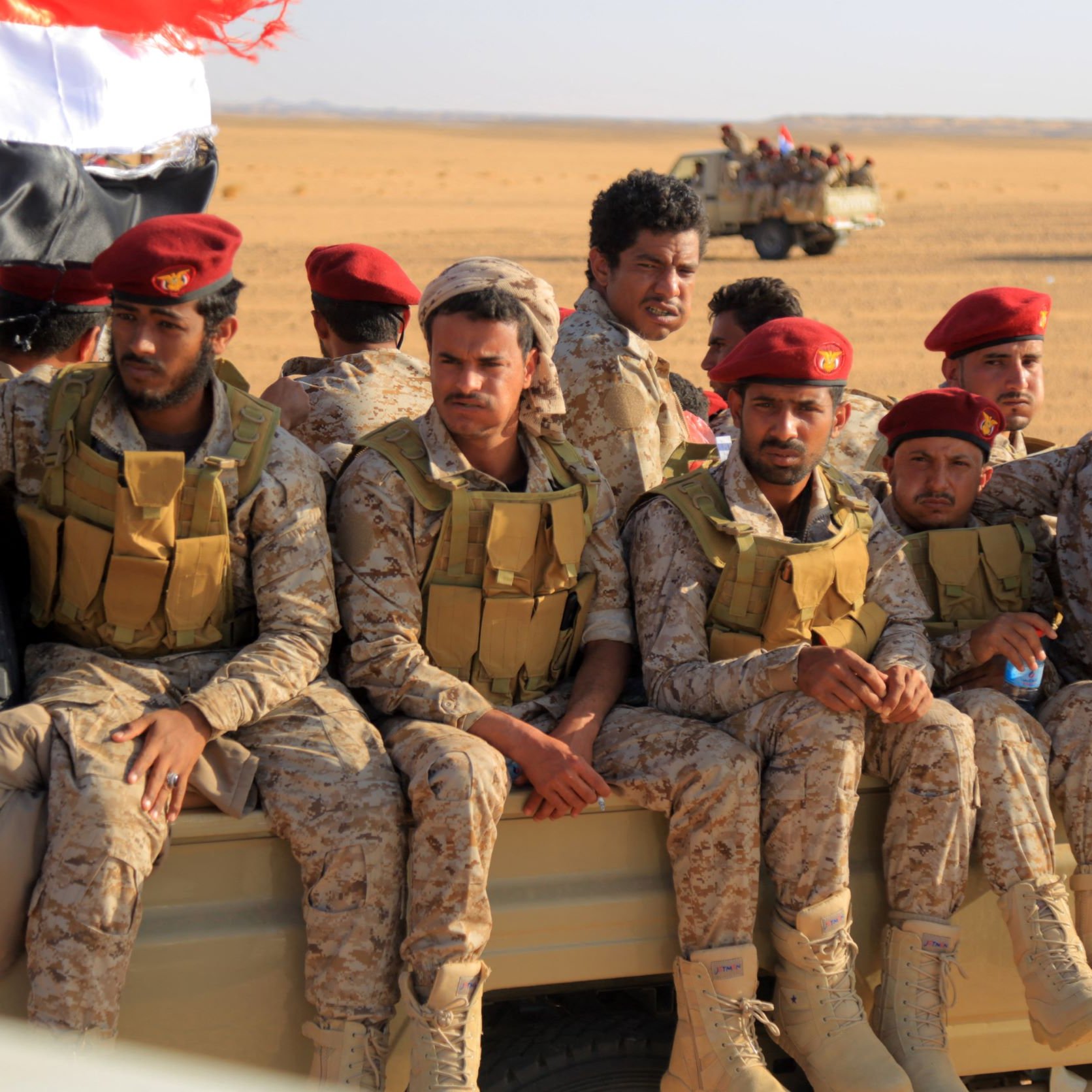 L'image montre un groupe de soldats assis à l'arrière d'un véhicule militaire dans un environnement désertique. Ils portent des uniformes camouflés et des casques ou des bérets rouges. À l'arrière-plan, on aperçoit d'autres véhicules militaires. Le paysage est aride et sablonneux, typique des zones désertiques. Les soldats semblent concentrés et disposés à exécuter leur mission.