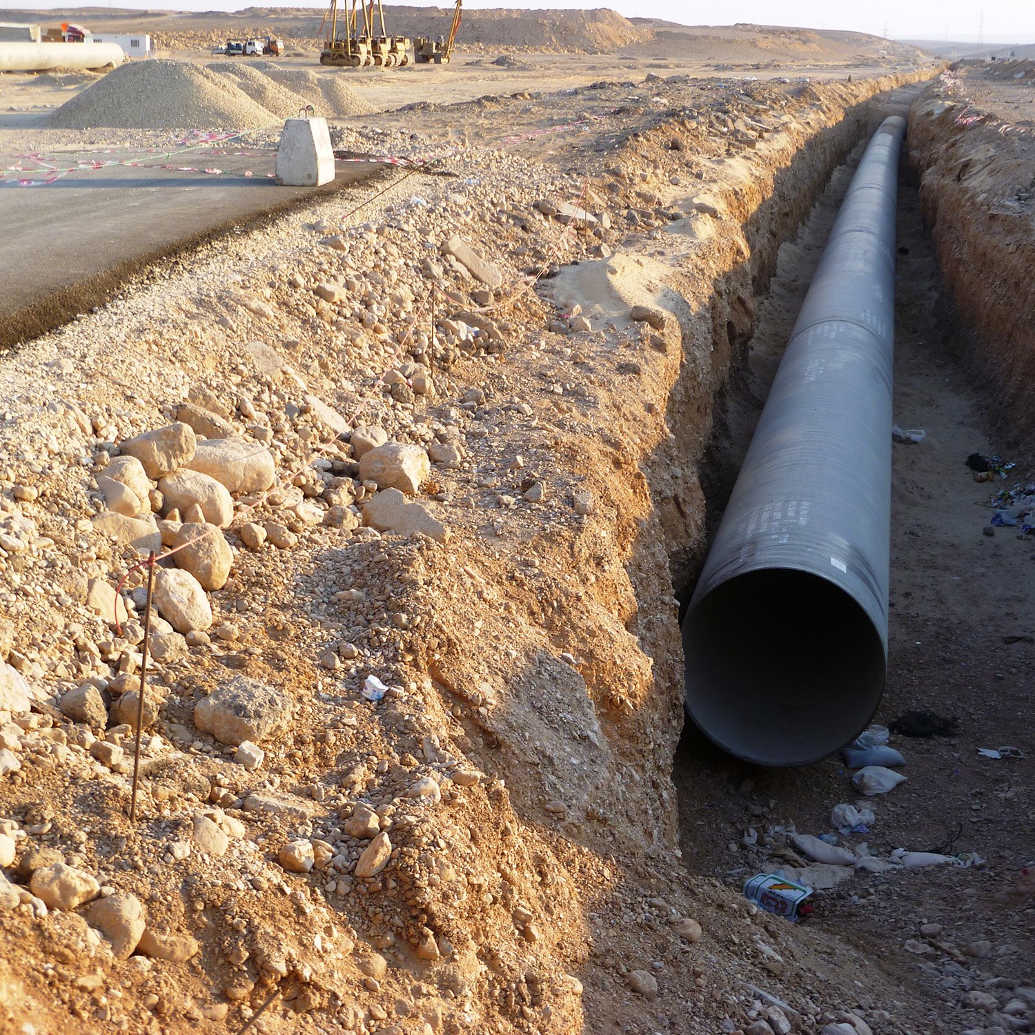 L'image montre un espace de construction où un grand tuyau en métal est disposé dans une tranchée creusée dans le sol. Le terrain environnant est aride et rocailleux, avec quelques débris, comme des bouteilles, dispersés dans la tranchée. Au fond, on peut voir des engins de chantier et une route partiellement construite. L'ambiance générale évoque un projet d'infrastructure en cours.