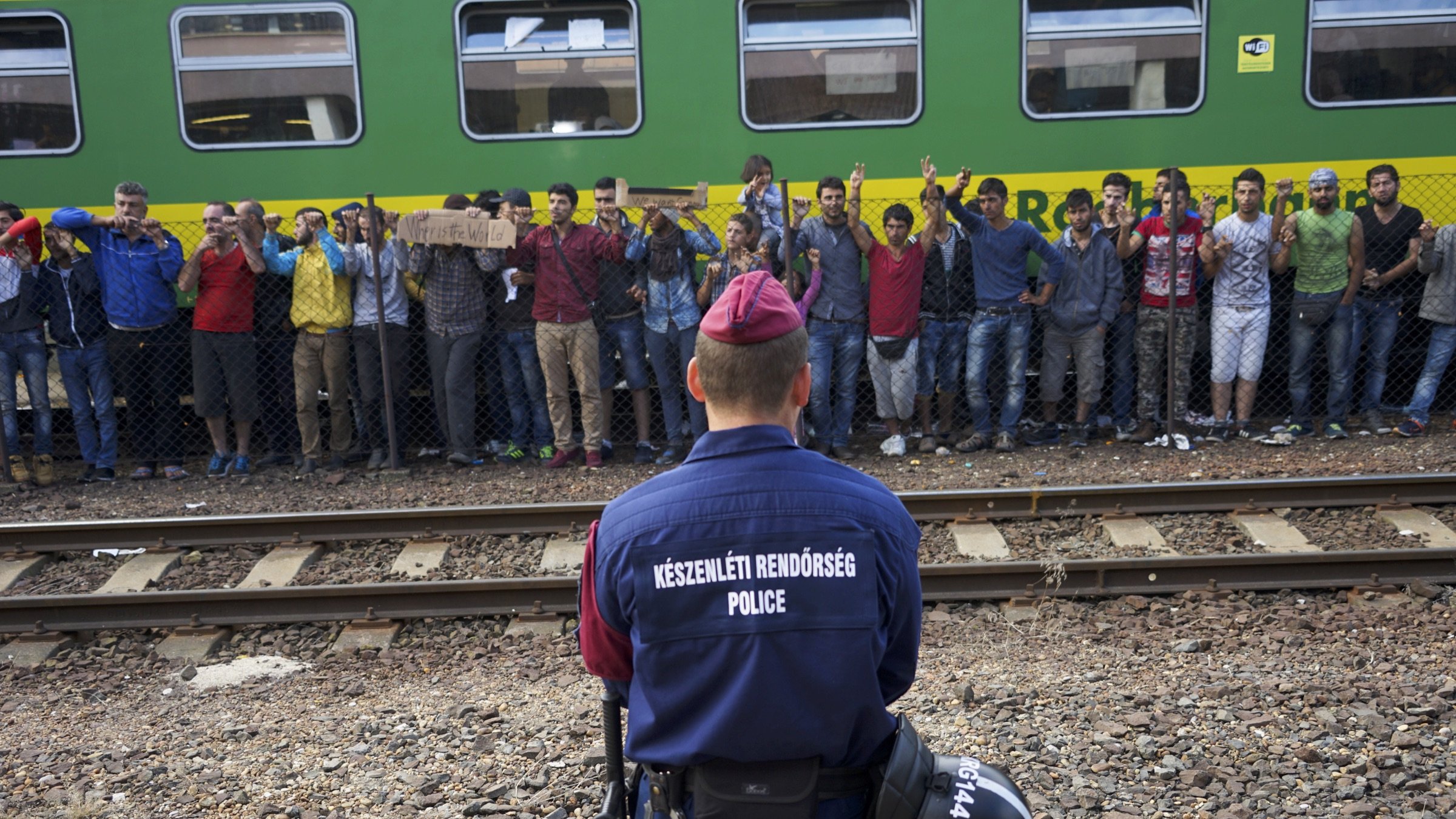 The image shows a scene at a train station where a police officer is facing a group of individuals. The officer is dressed in a uniform with the word "POLICE" clearly visible on the back. In front of the officer, there is a crowd of people, some holding signs, who appear to be expressing their discontent or making a statement. The background features a green train, and there are railway tracks visible, indicating that this is a transportation hub. The atmosphere seems tense, with the officer standing guard as the crowd raises their hands, possibly in protest or to seek attention.