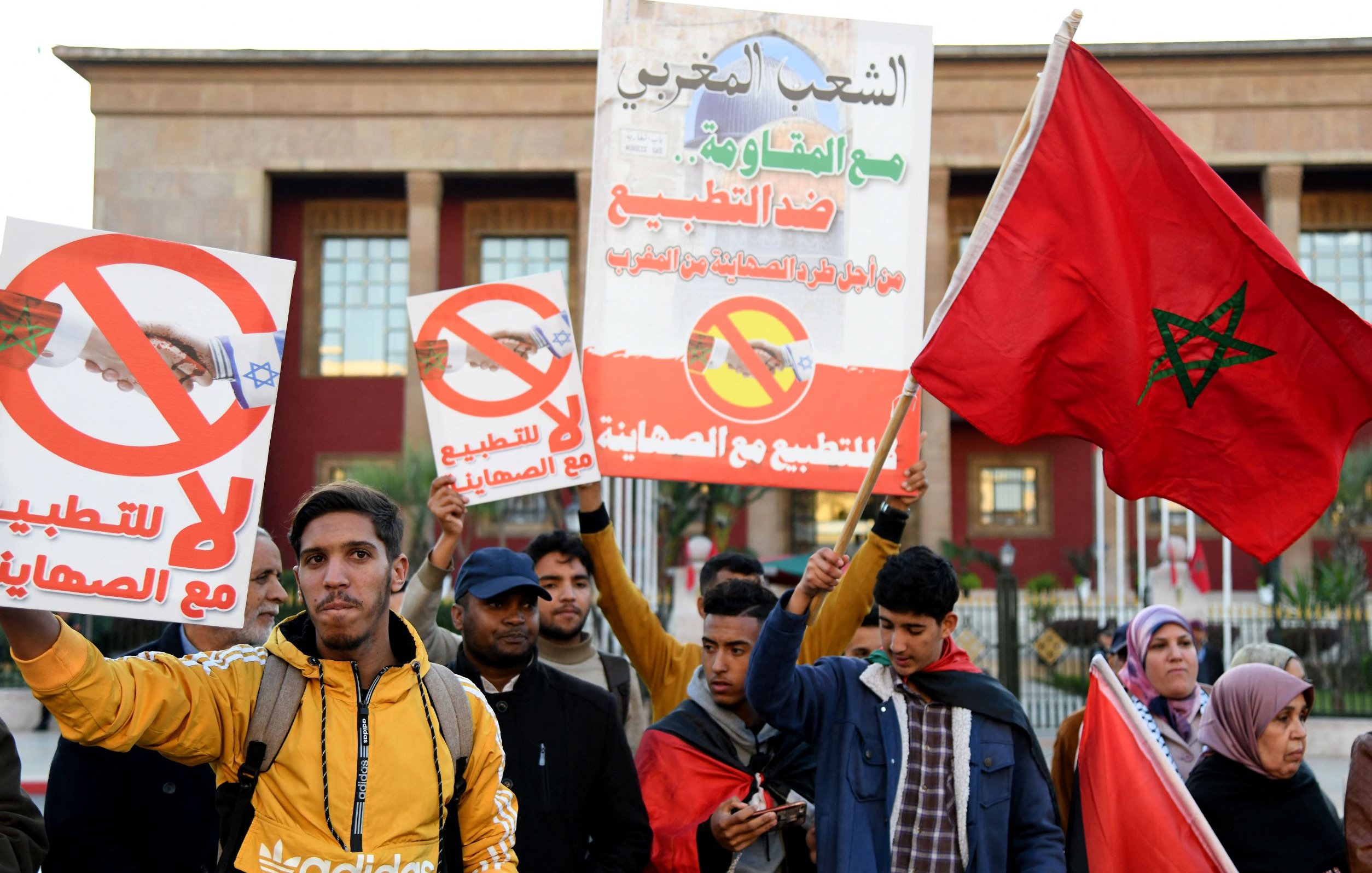 L'image montre un groupe de manifestants tenant des pancartes. Les affiches portent des messages visant à s'opposer à la normalisation des relations avec Israël, avec des symboles qui suggèrent un rejet de cette initiative. Les participants, d'âges divers, expriment leur désaccord à travers des slogans et des gestes. Certains portent des vêtements d'hiver, et l'ambiance semble être celle d'une manifestation pacifique. En arrière-plan, on aperçoit un bâtiment public, probablement lié au gouvernement.