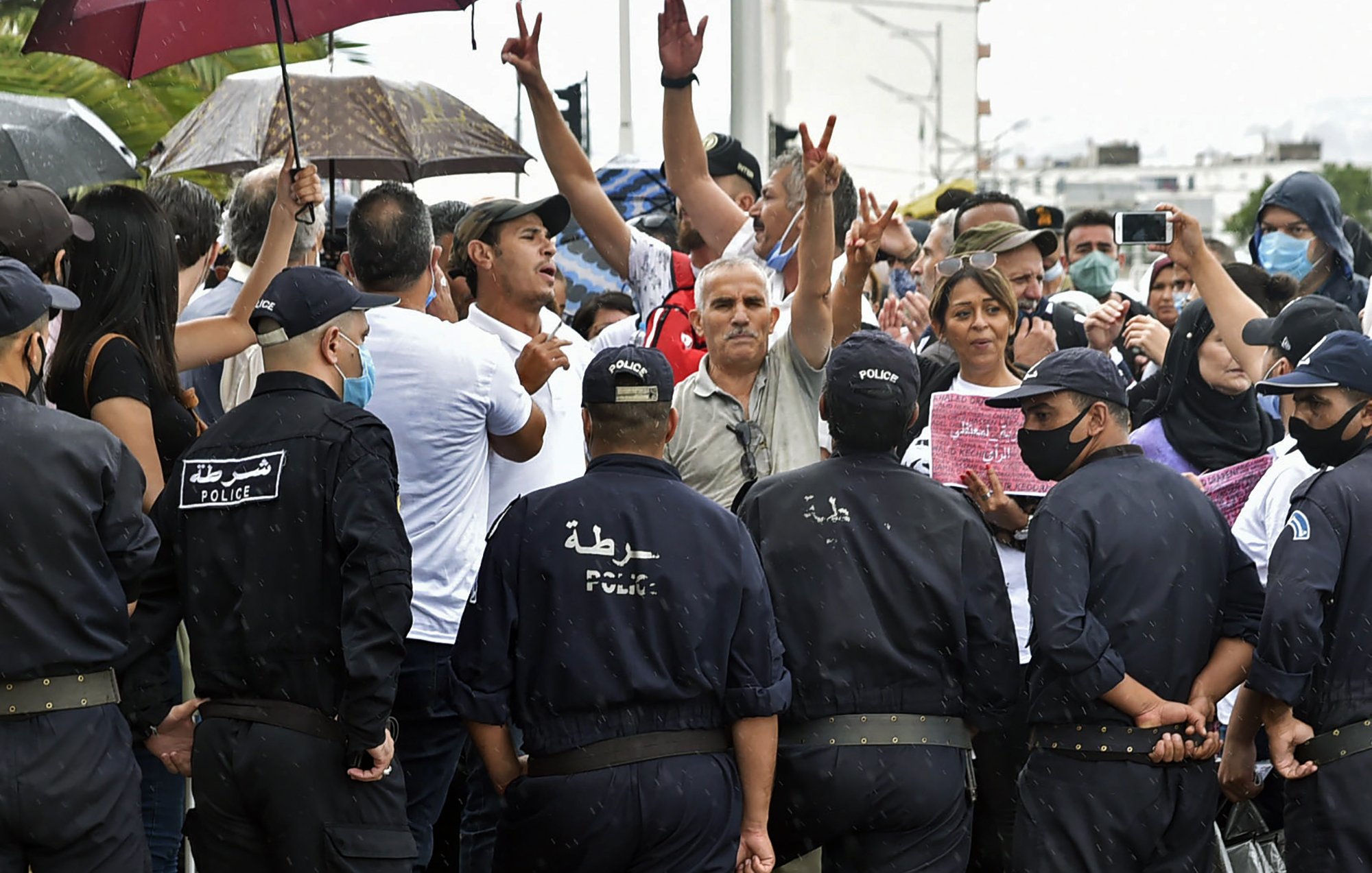 L'image montre une scène de tension entre un groupe de manifestants et des policiers. Les manifestants, certains avec des parapluies, lèvent les bras et expriment leurs opinions. Les policiers, en uniforme et portant des masques, se tiennent en face d'eux, créant un contraste clair entre les deux groupes. La situation semble chargée d'émotion, avec des personnes qui cherchent à faire entendre leur voix. Le temps est pluvieux, ajoutant une atmosphère dramatique à l'événement.