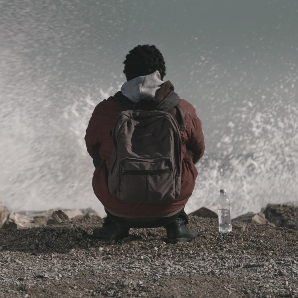 L'image montre une personne assise sur un rivage, tournant le dos à la caméra. Elle porte une veste et un sac à dos. À côté d'elle, il y a une bouteille d'eau. On peut voir des vagues qui se brisent sur les rochers, créant un effet de mouvement dans l'eau. L'atmosphère semble calme mais dynamique en raison du mouvement des vagues.