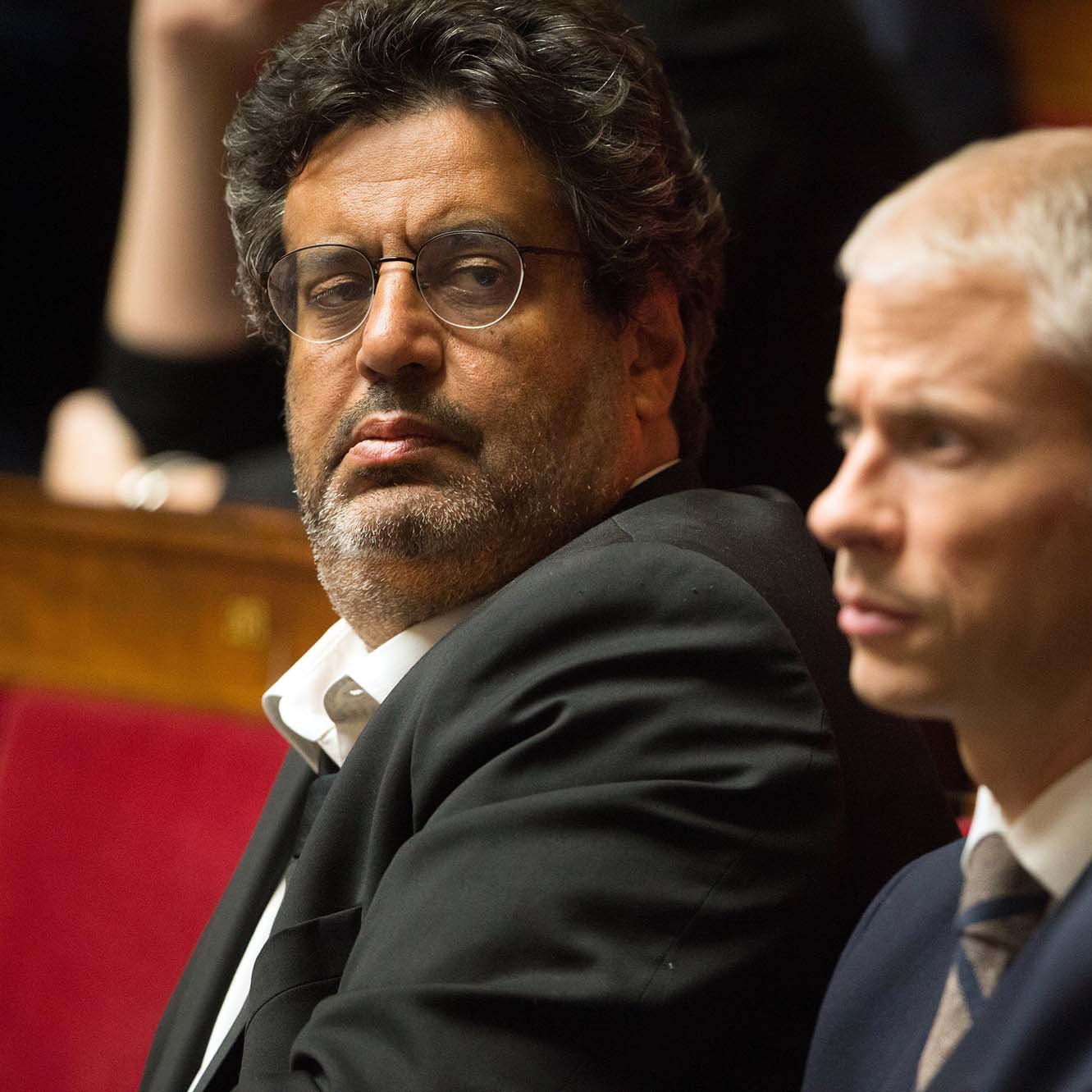 L'image montre un groupe d'hommes assis dans un cadre formel, probablement lors d'une séance législative. Un homme au premier plan porte des lunettes et un costume sombre, affichant une expression sérieuse, tandis que les autres à ses côtés semblent également attentifs et concentrés. L'arrière-plan montre des personnes floues, suggérant une ambiance politique. Les sièges sont ornés de rouge, typiques des assemblées.