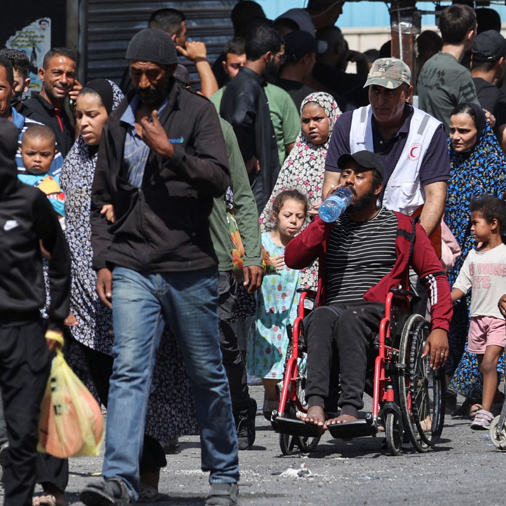 L'immagine mostra una scena affollata in un contesto urbano. Ci sono diverse persone che camminano insieme, tra cui adulti, bambini e alcune persone in carrozzina. Molte persone indossano abiti tradizionali o casual. C'è un'atmosfera vivace, ma al contempo seria, e i volti esprimono diverse emozioni. In primo piano, una persona in carrozzina tiene in mano una bottiglia d'acqua. Il contesto potrebbe suggerire una comunità che si riunisce, ma le condizioni dell'ambiente attorno sono visibili, con segni di spostamenti e attività quotidiane.