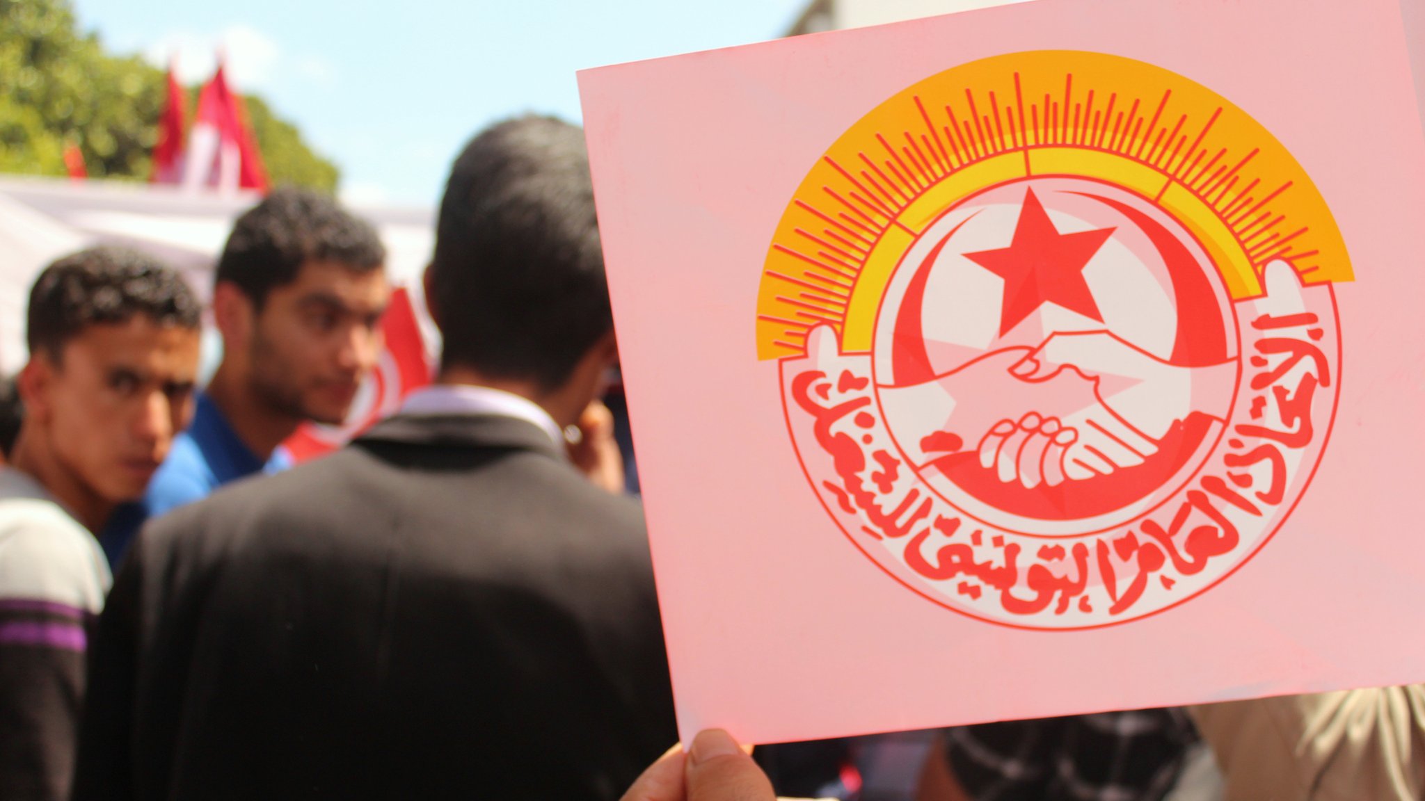 The image features a group of people, with a focus on one individual holding up a sign. The sign prominently displays a logo that includes two hands shaking, surrounded by a bright sunburst and red star. The text on the sign is in Arabic, likely associated with a labor or union organization. The background suggests an outdoor setting, possibly during a rally or demonstration, with other individuals visible nearby.