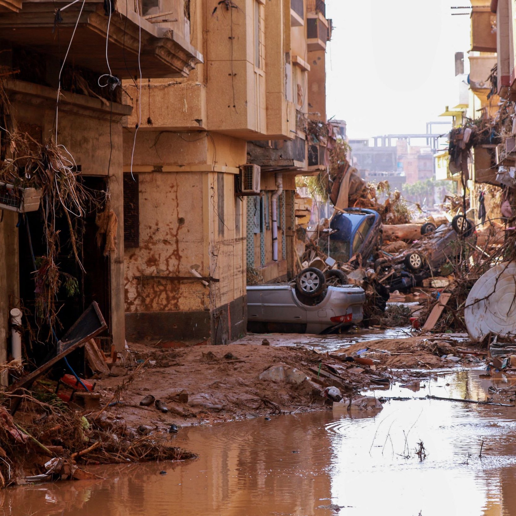 L'image montre une rue dévastée par une inondation. On peut voir des bâtiments endommagés, avec des débris et de la boue accumulés sur le sol. Des voitures sont renversées et se trouvent parmi les débris, tandis qu'une faible couche d'eau stagnante reflète la destruction environnante. L'atmosphère est sombre, soulignant les conséquences dramatiques de la catastrophe.