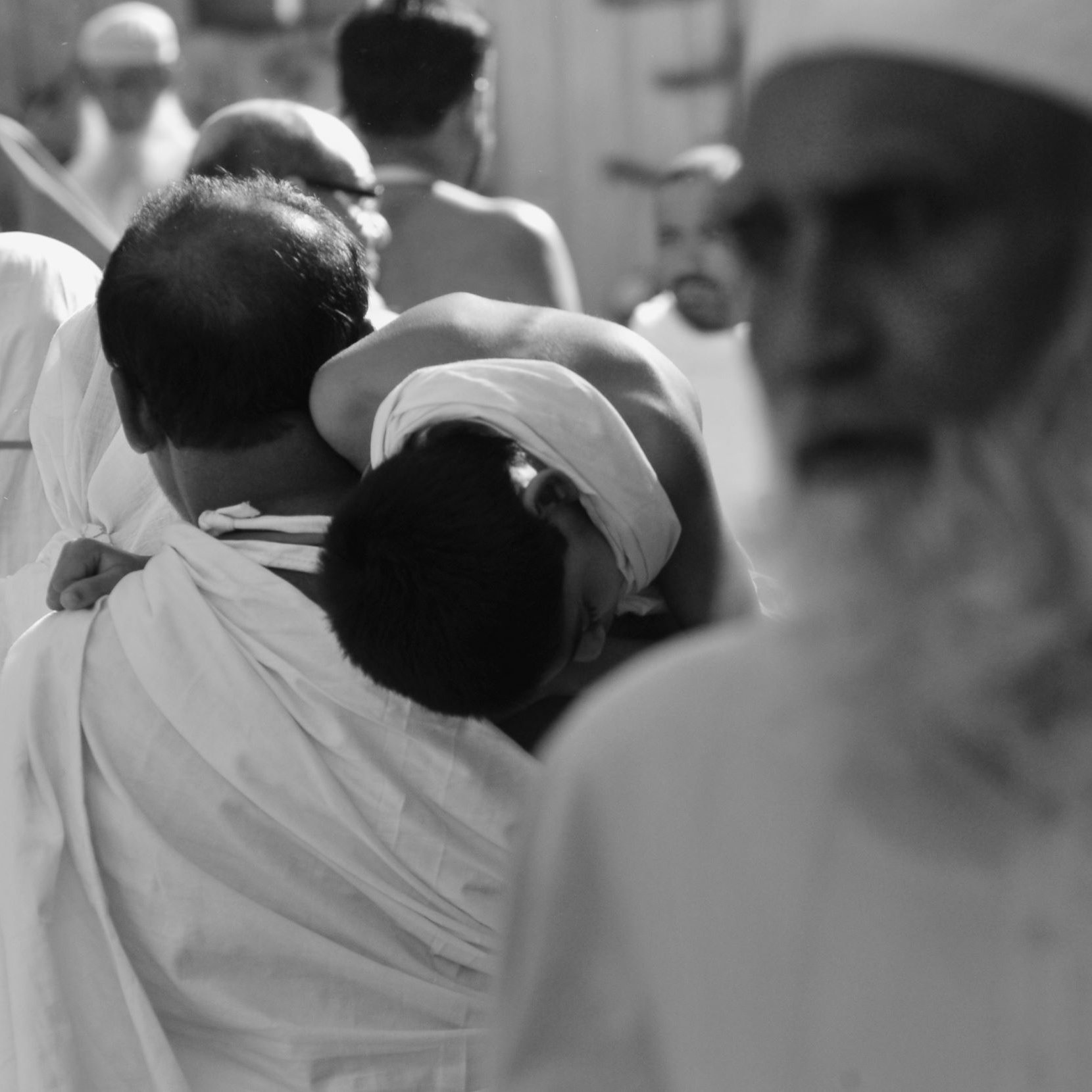 L'image montre une scène animée d'une foule, probablement lors d'un événement religieux. On peut voir un homme portant un enfant sur son dos, tandis que d'autres personnes sont visibles en arrière-plan, certaines portant des vêtements traditionnels. L'image est en noir et blanc, ce qui accentue l'atmosphère solennelle de l'événement. Les expressions des visages et les attitudes des personnes semblent refléter une ambiance de dévotion et de spiritualité.