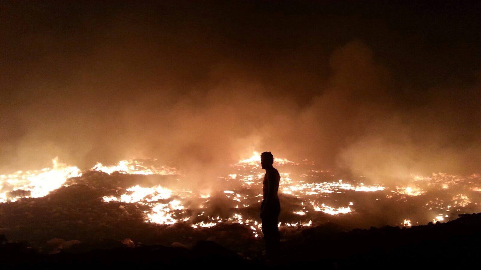 L'image montre un paysage de feu et de fumée. On voit un homme de silhouette, se tenant debout au premier plan, face à un vaste champ d'incendie. Le sol est enflammé, et une épaisse fumée s'élève, créant une atmosphère sombre et chaotique. Les flammes et la lumière orangée contrastent avec l'obscurité environnante, soulignant la gravité de la situation.