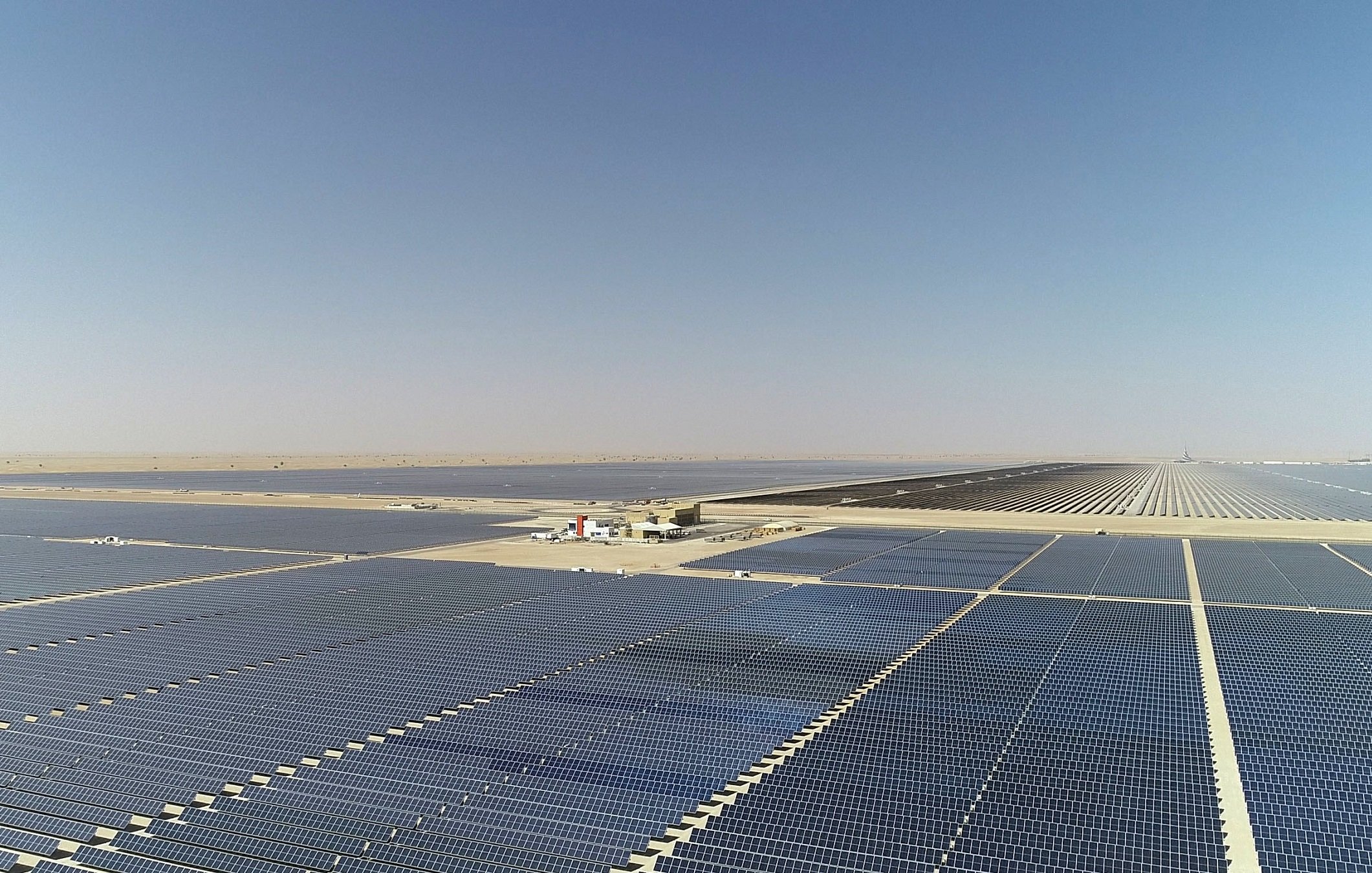 تظهر الصورة مزرعة شمسية كبيرة تحتوي على عدد كبير من الألواح الشمسية المنتشرة على مساحة واسعة. الألواح مرتبة في صفوف متوازية وتمتد إلى الأفق تحت سماء صافية وخالية من الغيوم. في الخلفية، يمكن رؤية محطة توليد الطاقة أو منشأة صغيرة تابعة للمزرعة، مما يدل على استغلال الطاقة الشمسية في توليد الكهرباء. تعكس الألواح الشمسية أشعة الشمس، مما يبرز نطاق المشروع وأهميته كجزء من استراتيجيات الطاقة المتجددة.