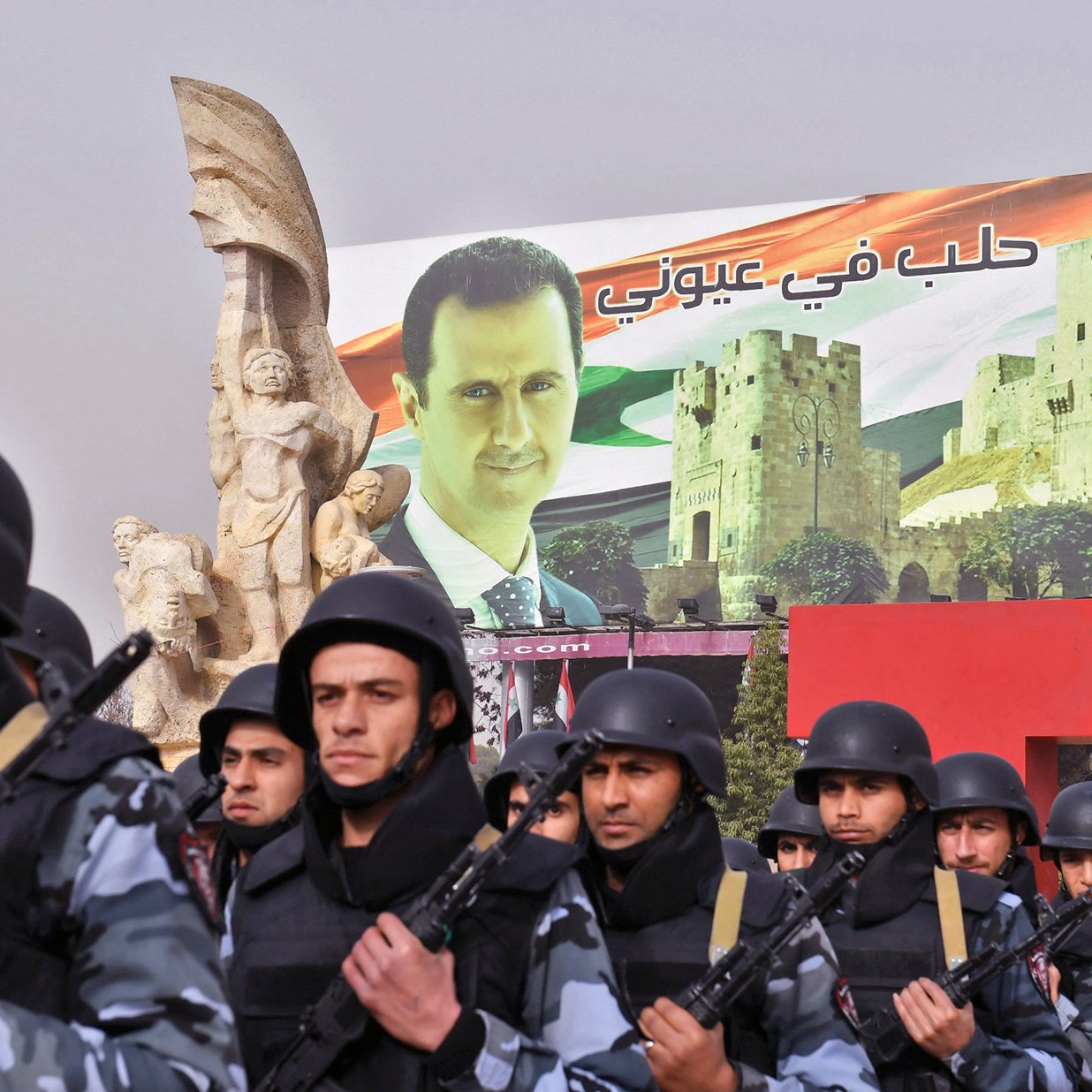 تظهر الصورة عرضًا عسكريًا أو تظاهرة، حيث يسير مجموعة من الجنود في صفوف منظمة، يرتدون زيًا عسكريًا ويحمون أسلحة. خلفهم يوجد لافتة ضخمة تحمل صورة لرجل، تترافق مع مشاهد معمارية تتعلق بسوريا. تظهر أيضًا بعض الرموز الوطنية مثل الأعلام. الأجواء تعكس نوعًا من القوة والتأييد السياسي.