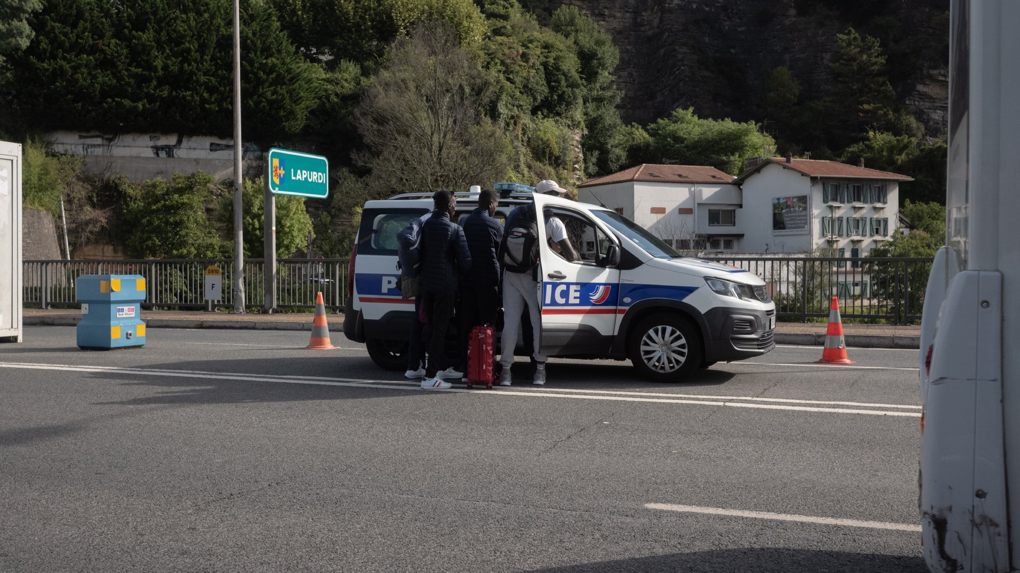 L'image montre une scène sur une route, où un véhicule de police est stationné. Des personnes se tiennent près de la voiture de police, probablement en train de discuter avec les agents. En arrière-plan, on peut voir quelques bâtiments et de la végétation. Des cônes de signalisation sont visibles sur la voie, indiquant peut-être une opération de contrôle ou de sécurité.