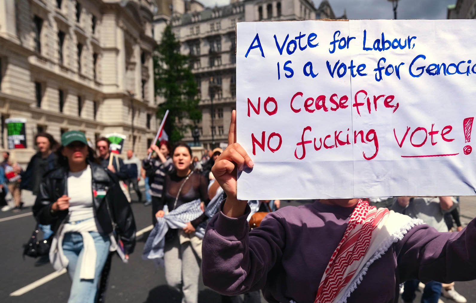 تصویر مربوط به یک تظاهرات است. در جلوی تصویر فردی دیده می‌شود که تابلویی را در دست دارد. روی تابلو نوشته شده: "رای به کارگر، رای به نسل‌کشی است" و همچنین جمله‌ای دیگر به این مضمون که "نه آتش‌بس، نه رای لعنتی!" در پس‌زمینه، جمعیتی از معترضان در حال راهپیمایی دیده می‌شوند. فضای عمومی و شهری است و مردم با شور و هیجان در حال اظهار نظر در مورد موضوعات اجتماعی و سیاسی هستند.