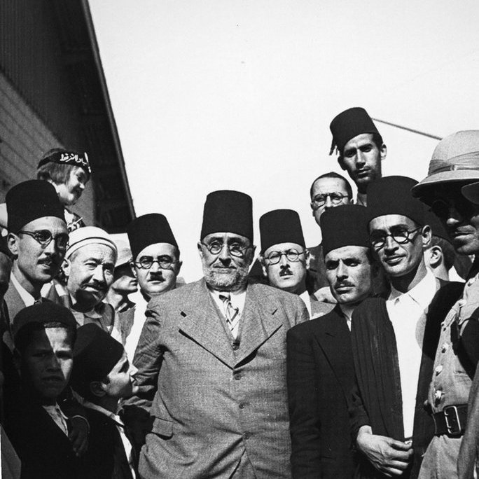 L'image représente un groupe d'hommes en costumes formels, majoritairement portant des fez. Ils semblent être engagés dans une discussion ou un moment de camaraderie. L'ambiance est sérieuse, et les expressions des visages montrent une concentration ou une détermination. Une femme est également visible en arrière-plan, portant un foulard. Le cadre pourrait indiquer un événement historique ou un rassemblement important, probablement dans un contexte culturel ou politique.