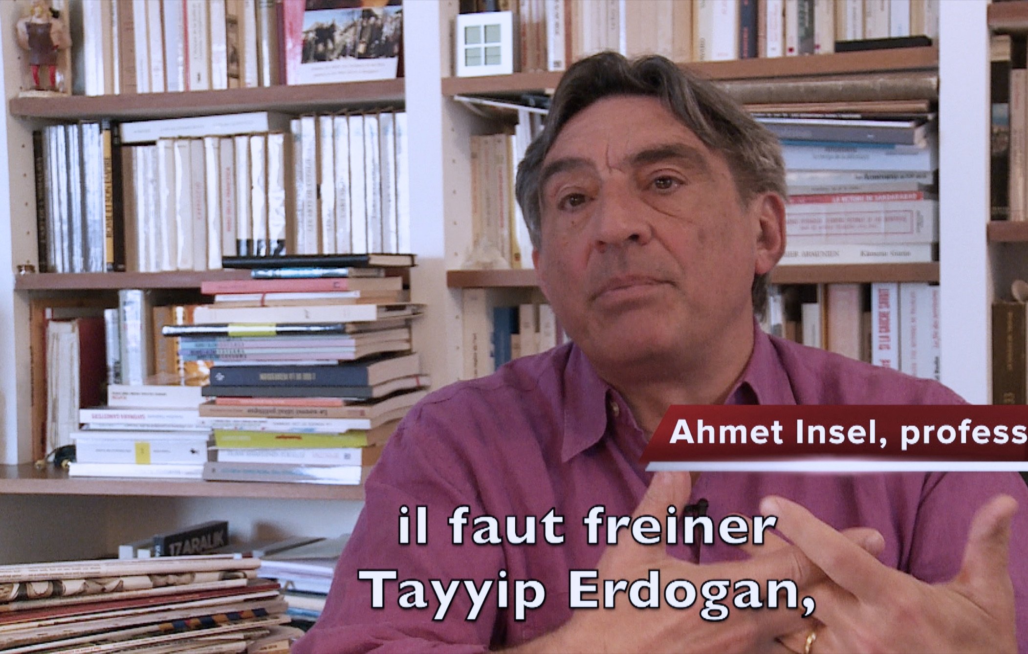 L'image montre un homme assis dans un environnement intérieur, entouré de livres et de dossiers sur des étagères. Il semble s'adresser à un interlocuteur ou à une caméra. L'homme est identifié comme Ahmet Insel, un professeur, et il exprime une opinion sur la nécessité de freiner Tayyip Erdogan. Il porte une chemise de couleur mauve et utilise des gestes pour appuyer ses propos. Les livres en arrière-plan suggèrent un cadre intellectuel.