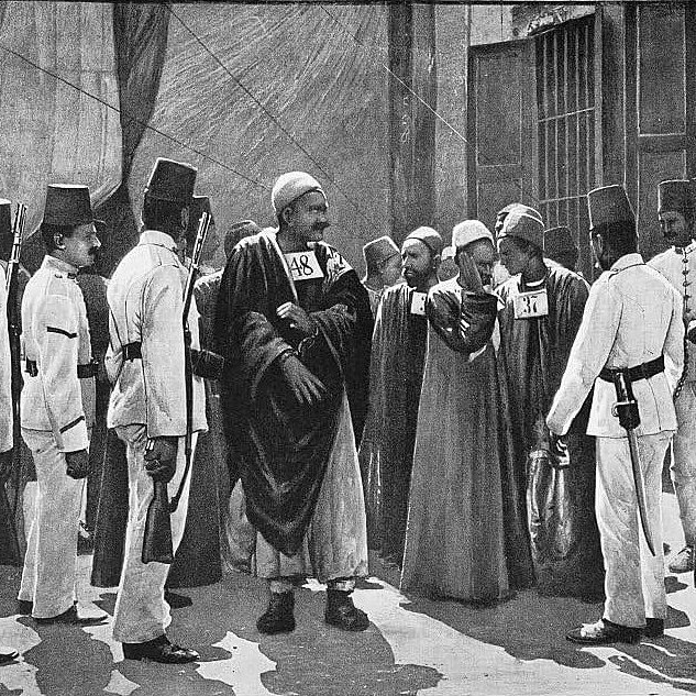 تظهر الصورة مجموعة من الأشخاص في سياق تاريخي، حيث يرتدي بعضهم زيًا تقليديًا يتضمن طربوشًا، بينما يرتدي آخرون زيًا رسميًا أو عسكريًا. يبدو أنهم في موقف اجتماعي أو رسمي، حيث يتبادلون الحديث أو يتفاوضون. يمكن ملاحظة الأرقام المدونة على صدور بعض الأشخاص، مما يشير إلى نظام ما أو ترتيب معين. الخلفية تبدو بسيطة، مما يركز الانتباه على الأشخاص ومكان تواجدهم.