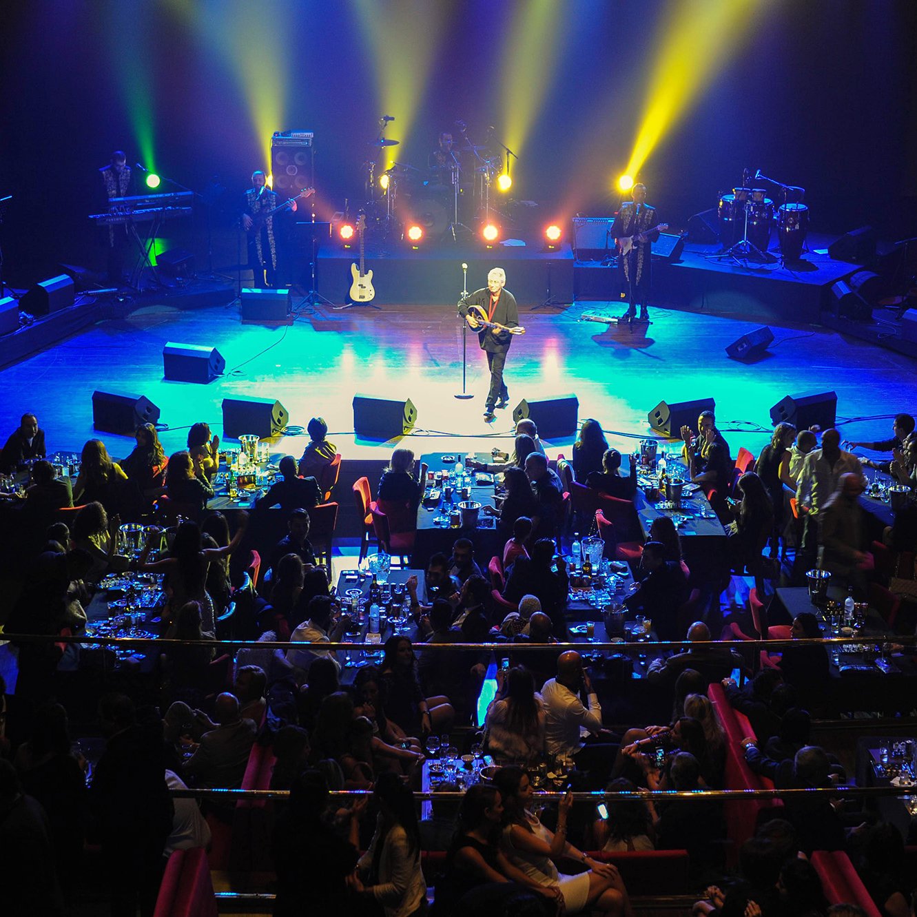 L'image montre une scène de concert animée. Au premier plan, un artiste se produit sur scène, entouré de lumières colorées et de spots éclairant l'espace. En arrière-plan, on peut voir des musiciens qui l'accompagnent. La salle est pleine de spectateurs assis à des tables, savourant leur soirée avec des repas et des boissons. L'ambiance est festive et chaleureuse, avec des gens qui semblent apprécier la performance.