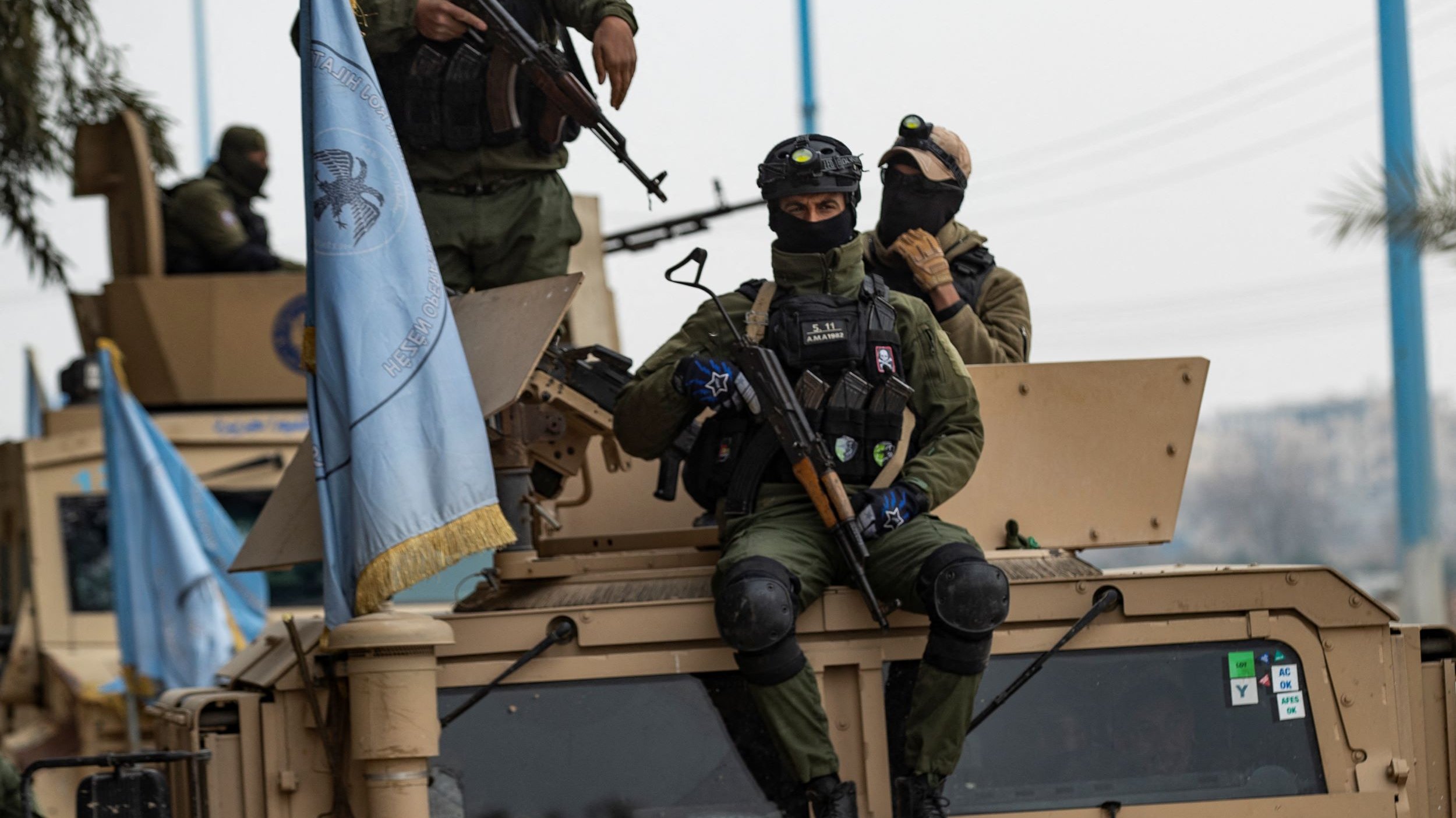 L'image montre des soldats en uniforme, armés et équipés de casques, se trouvant sur un véhicule militaire. L'un des soldats est assis sur le toit du véhicule, tenant une arme, tandis que d'autres soldats sont debout à proximité. On peut également voir un drapeau, probablement lié à une mission de maintien de la paix. L'environnement semble urbain, avec des éléments de la ville en arrière-plan.