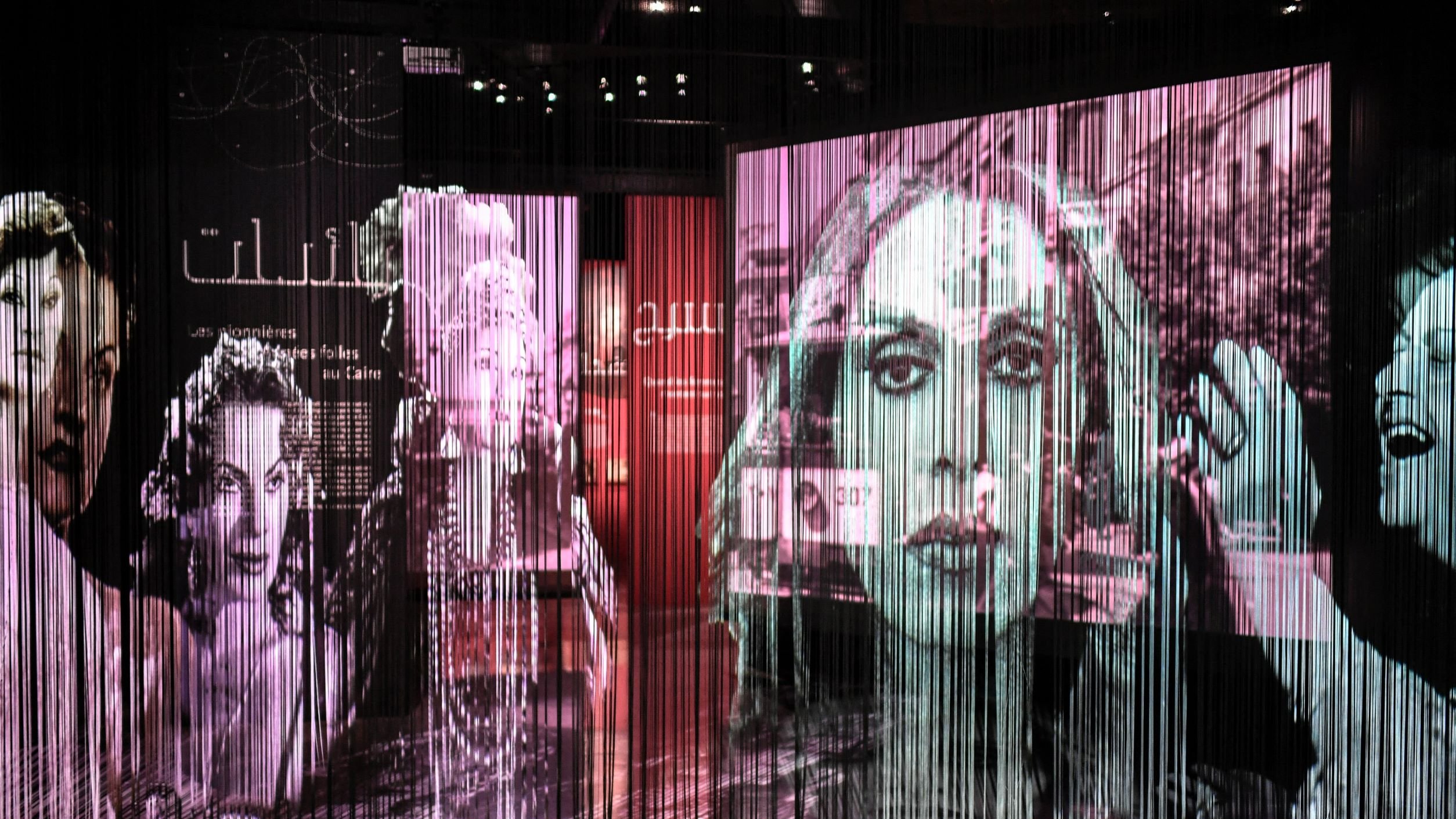 L'immagine presenta una serie di proiezioni artistiche su uno sfondo di tende di tessuto trasparente. Le immagini ritraggono volti femminili e scene di vita, con un forte contrasto tra bianco e nero e tonalità colorate. L'atmosfera è suggestiva e intimista, creando un effetto di profondità e movimento. Le fotografie sembrano evocare un senso di nostalgia, magari legato alla storia e alla cultura. Le scritte in arabo e altre lingue suggeriscono un tema culturale e un messaggio profondo.