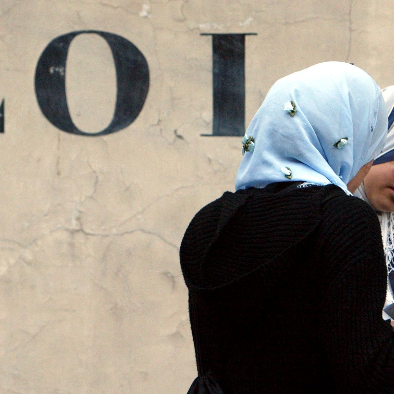 L'image présente deux femmes discutant, l'une d'elles portant un hijab bleu clair et l'autre un hijab sombre. Elles semblent concentrées sur un document ou une feuille qu'elles regardent ensemble. En arrière-plan, on peut voir le mot "LOI" écrit en lettres majuscules sur un mur beige, ce qui donne un contexte à la scène. L'atmosphère semble calme et introspective.