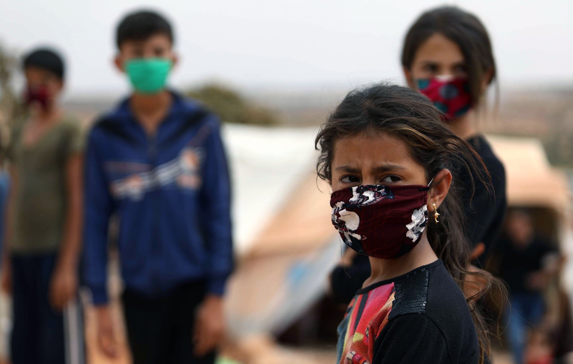 الصورة تظهر مجموعة من الأطفال في مخيم، حيث يرتدي بعضهم أقنعة واقية. في المقدمة توجد فتاة صغيرة تبدو قلقة، بينما خلفها يعبر الأطفال عن مشاعر متفاوتة. الخلفية غير واضحة، مما يشير إلى أنهم في بيئة مفتوحة لكنها غير مريحة. الإضاءة تبدو طبيعية، مما يعكس واقع الحياة اليومية في المخيم.