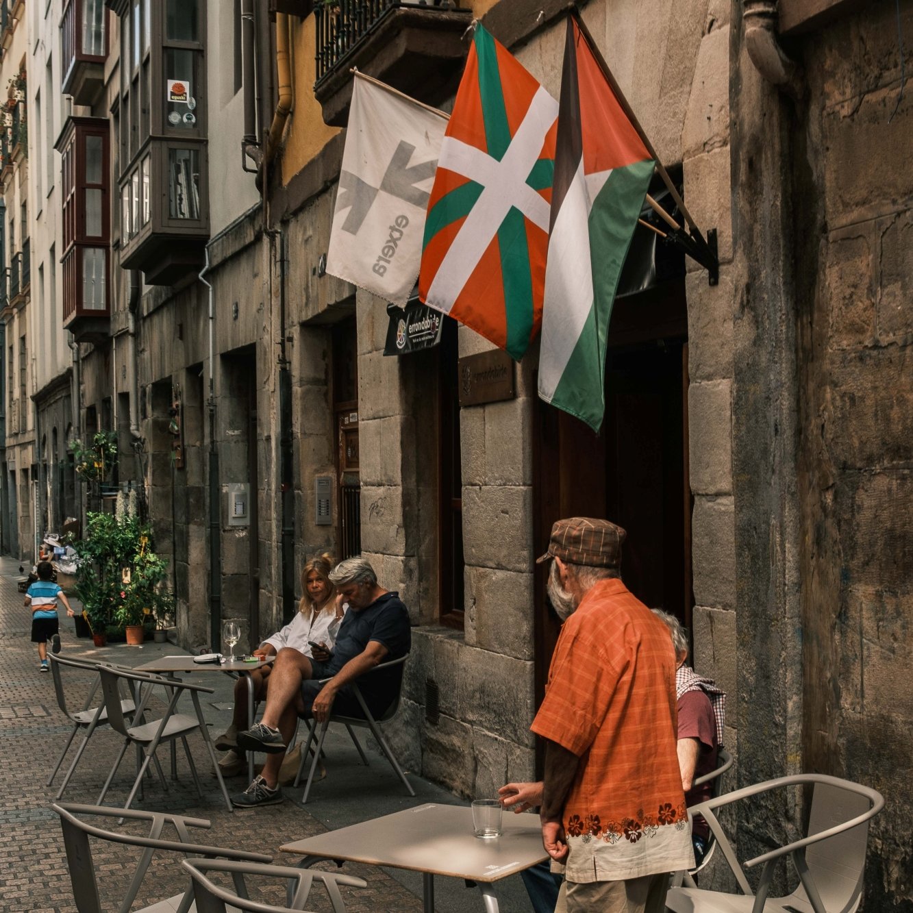Ruelle animée avec tables de café, drapeaux et passants au style basque.