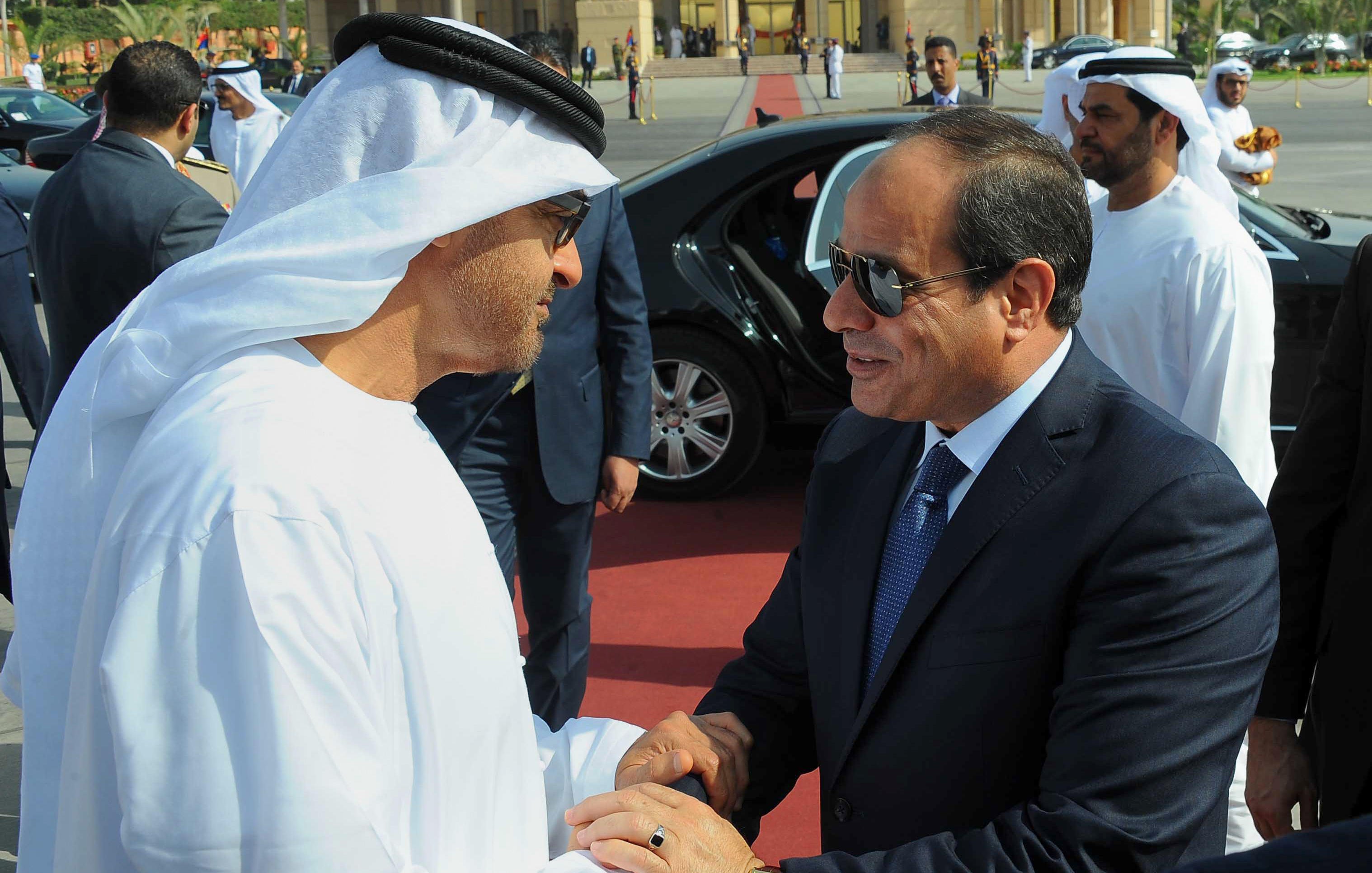L'image montre une rencontre entre deux dirigeants, probablement en train de se saluer avec une poignée de main. Les individus sont vêtus de vêtements traditionnels, comme des kandouras et des keffiehs pour l'un d'eux. On peut également voir d'autres personnes en arrière-plan, ainsi qu'une voiture haut de gamme, suggérant un événement officiel ou une cérémonie. L'ambiance semble cordiale et respectueuse.