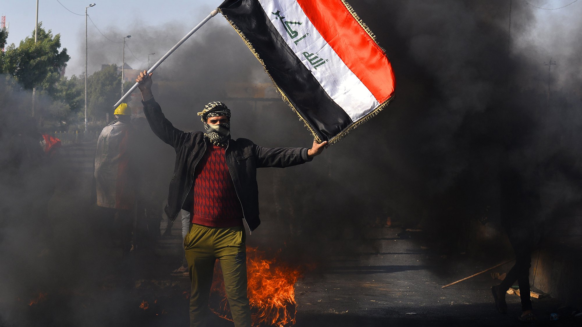 The image depicts a person holding the Iraqi flag amidst a scene of protest. The individual is wearing a mask and has their face partially covered, standing in front of billowing black smoke and flames. The background suggests a chaotic environment, likely indicative of unrest or demonstrations. The focus on the flag emphasizes national identity in the context of the protest.
