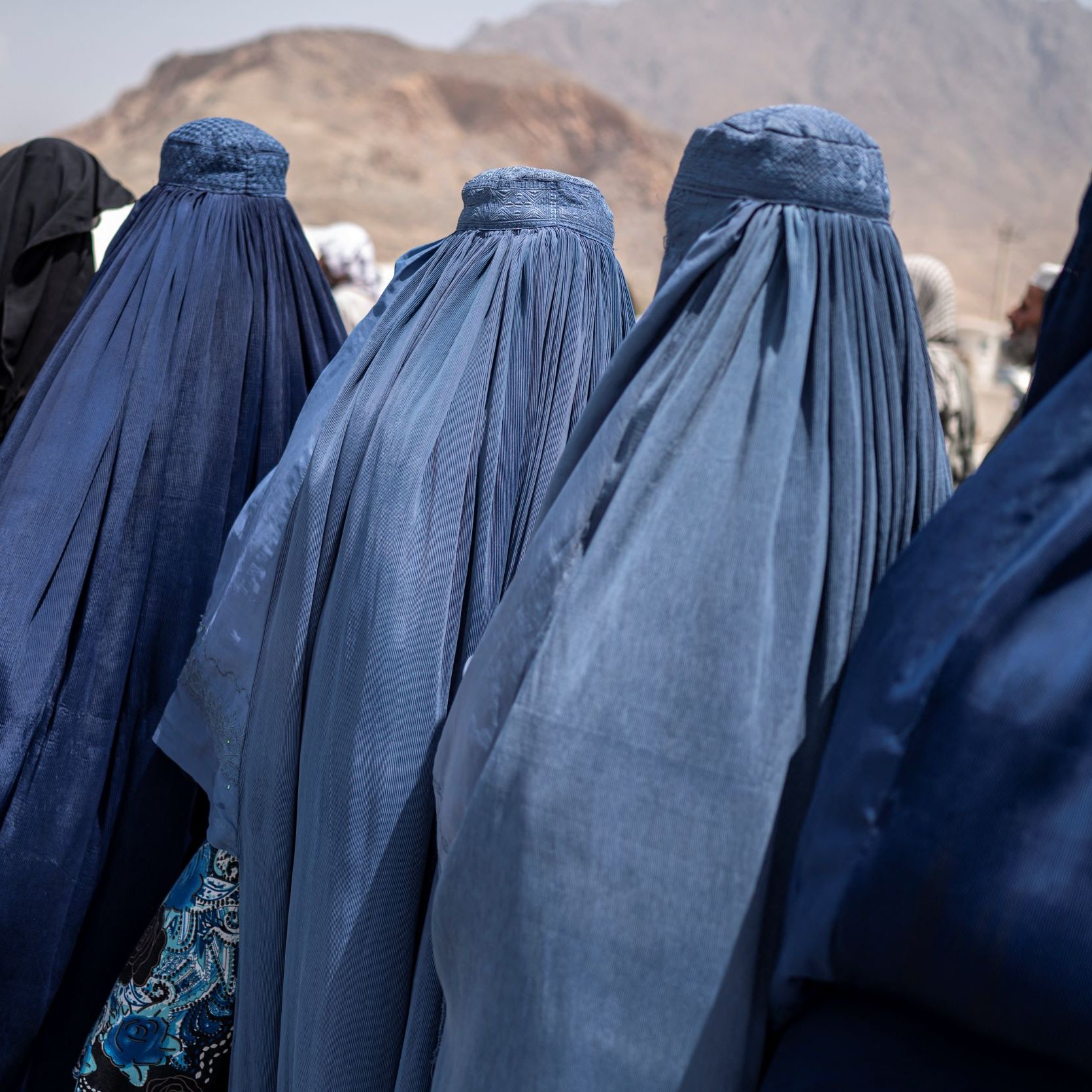 در این تصویر تعدادی زنان دیده می‌شوند که پوشش‌هایی به رنگ‌های آبی و مشکی بر تن دارند. آنها از پشت به دوربین ایستاده‌اند و فقط قسمت‌هایی از پوشش‌هایشان قابل مشاهده است. نمای背景 کوه‌ها و فضای باز است، که ممکن است به منطقه‌ای کوهستانی اشاره داشته باشد. این تصویر حس سنتی و فرهنگی خاصی را منتقل می‌کند.