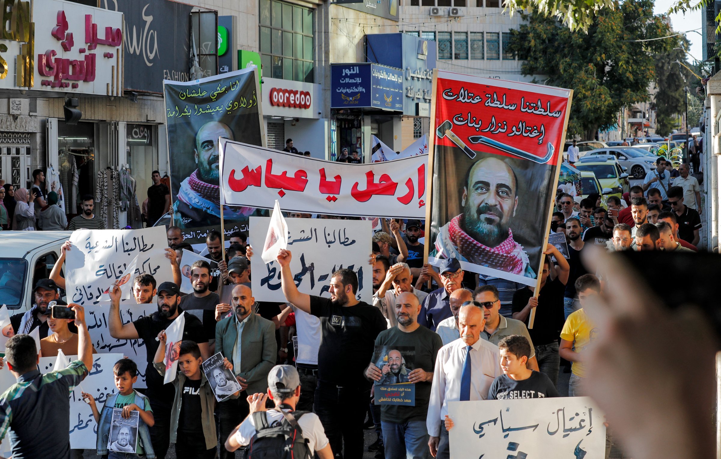 L'image montre une foule de manifestants dans une rue. Les participants portent des pancartes et des banderoles avec des écrits en arabe. L'ambiance semble être celle d'une manifestation ou d'un rassemblement public. Plusieurs personnes brandissent des affiches, et on peut voir des voitures et des bâtiments en arrière-plan. Les manifestants semblent engagés et déterminés dans leur action.