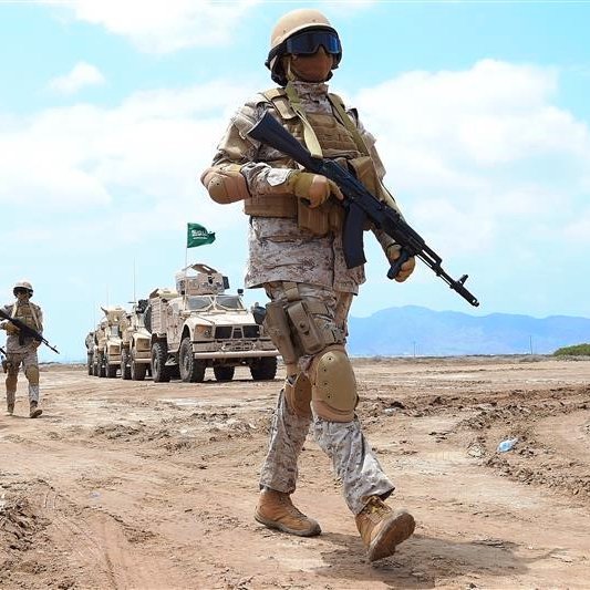 L'image montre des soldats en tenue militaire marchant sur un terrain désertique. Ils portent des casques et des gilets pare-balles, et certains utilisent des armes. En arrière-plan, on peut voir des véhicules militaires. Le ciel est clair, et le paysage est aride. La scène évoque une ambiance de formation ou d'exercice militaire.