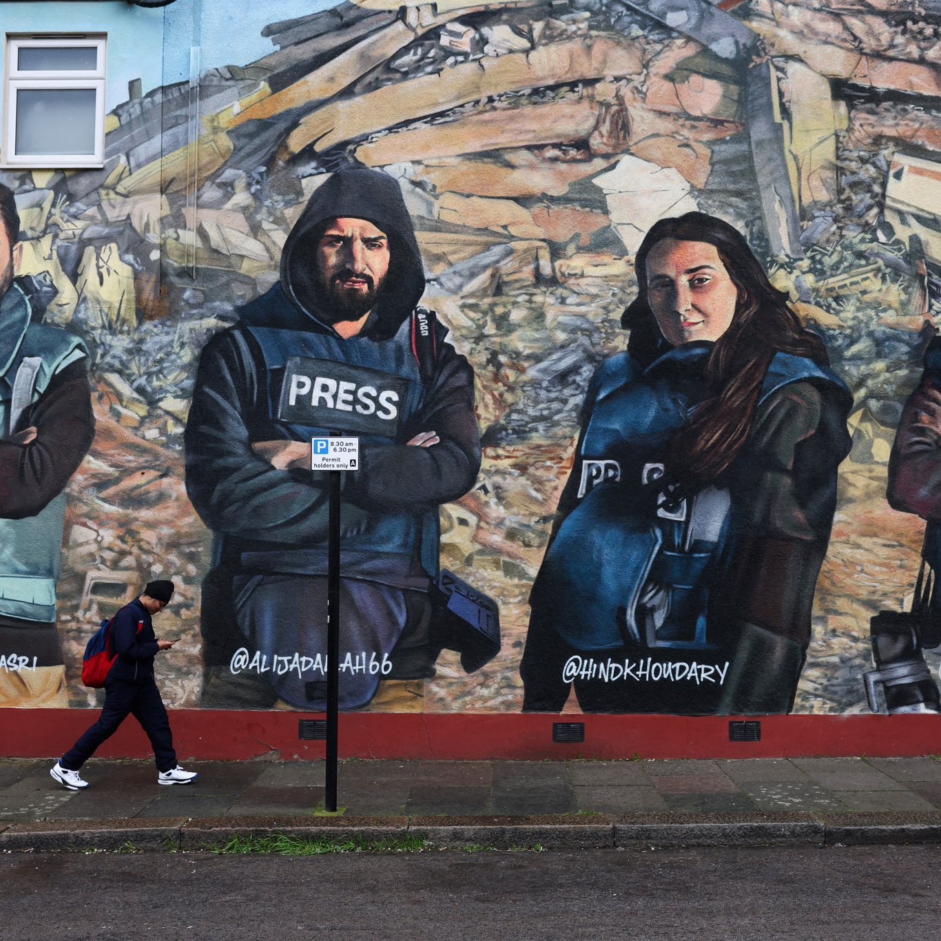L'image montre une fresque murale représentant quatre personnes habillées en gilets pressés, posant avec les bras croisés. Ils semblent symboliser des journalistes ou des reporters. En arrière-plan, il y a des débris, suggérant un contexte de destruction ou de conflit. Une personne marche au premier plan, ajoutant une dynamique à la scène. Les visages des individus sur la fresque sont sérieux et déterminés.