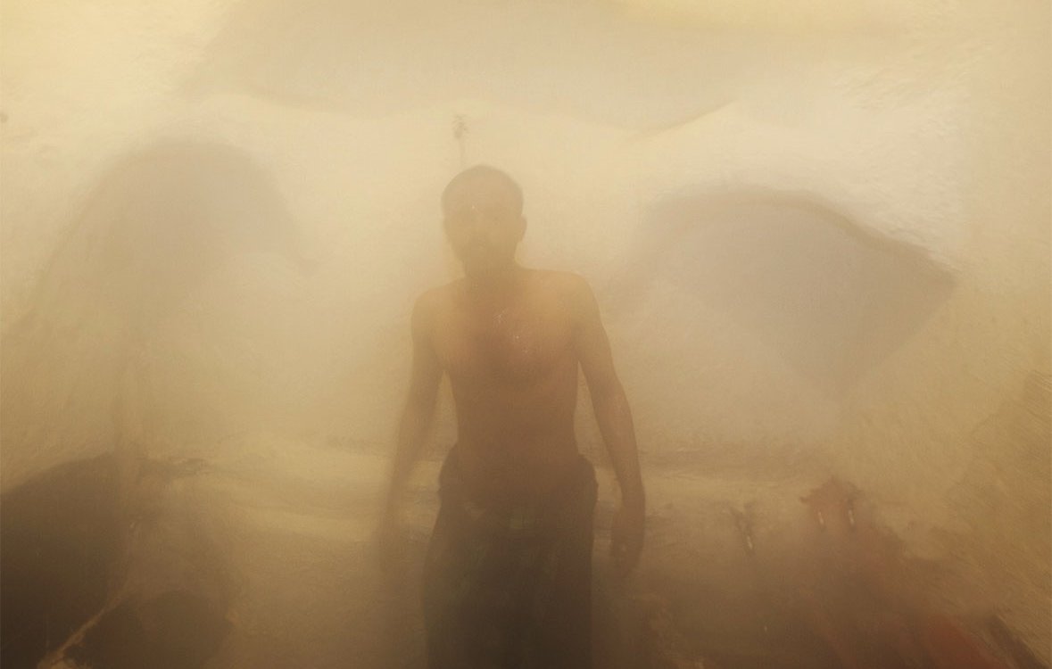 L'image montre un homme dans un environnement humide et brumeux, probablement à l'intérieur d'un hammam ou d'un sauna. La vapeur crée une atmosphère indistincte et floue, rendant les contours des objets difficilement visibles. L'homme est torse nu et semble se tenir debout, enveloppé par la vapeur qui l'entoure, ajoutant une sensation de chaleur et de sérénité à la scène. Les murs sont d'apparence texturée, typiques des espaces de bien-être.