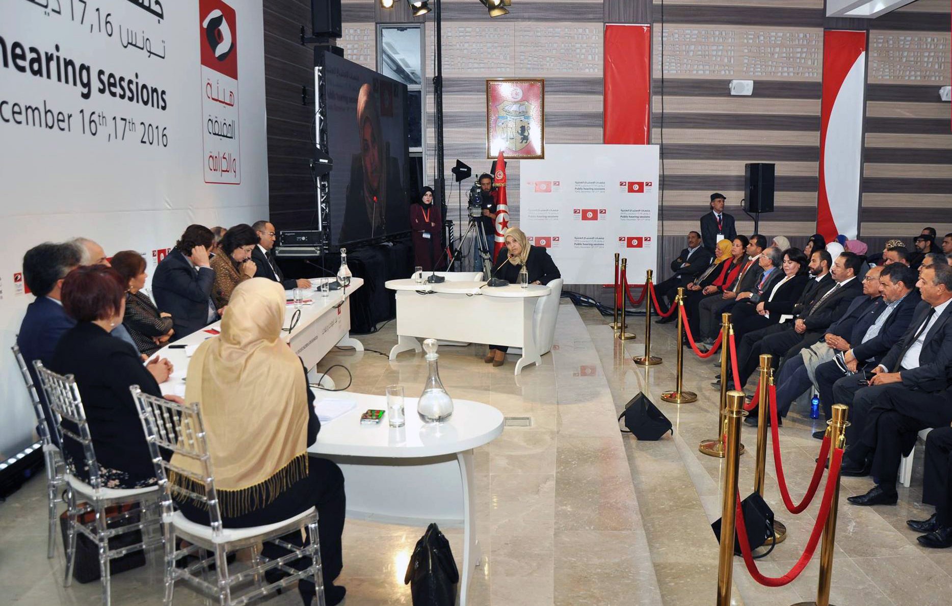 L'image montre un panel de discussion ou une session d'audition, avec plusieurs personnes assises face à une table à l'avant. Au fond, il y a une bannière affichant des informations sur l'événement, datée des 16 et 17 décembre 2016. Les participants semblent engagés dans la discussion, avec une variété de personnes, y compris des femmes portant des foulards. La scène est bien organisée, avec des cordons de sécurité délimitant l'espace où se trouvent le public et les intervenants.