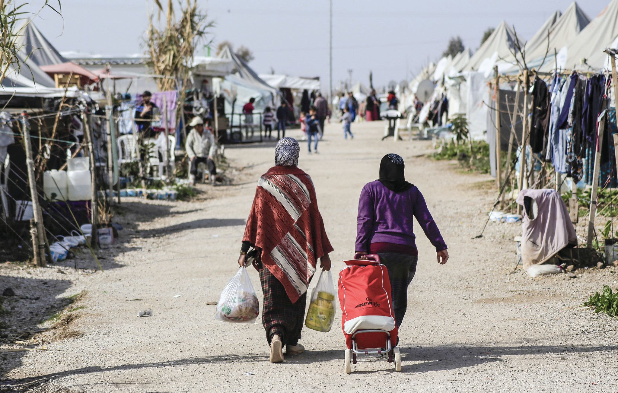 L'image montre une scène d'un camp, probablement un camp de réfugiés ou un site temporaire. Deux femmes marchent sur un chemin de terre, portant des sacs. L'une d'elles pousse une poussette. En arrière-plan, on aperçoit des tentes et des structures improvisées, avec d'autres personnes visibles dans l'environnement. L'atmosphère semble être celle d'une communauté, avec des éléments de vie quotidienne.