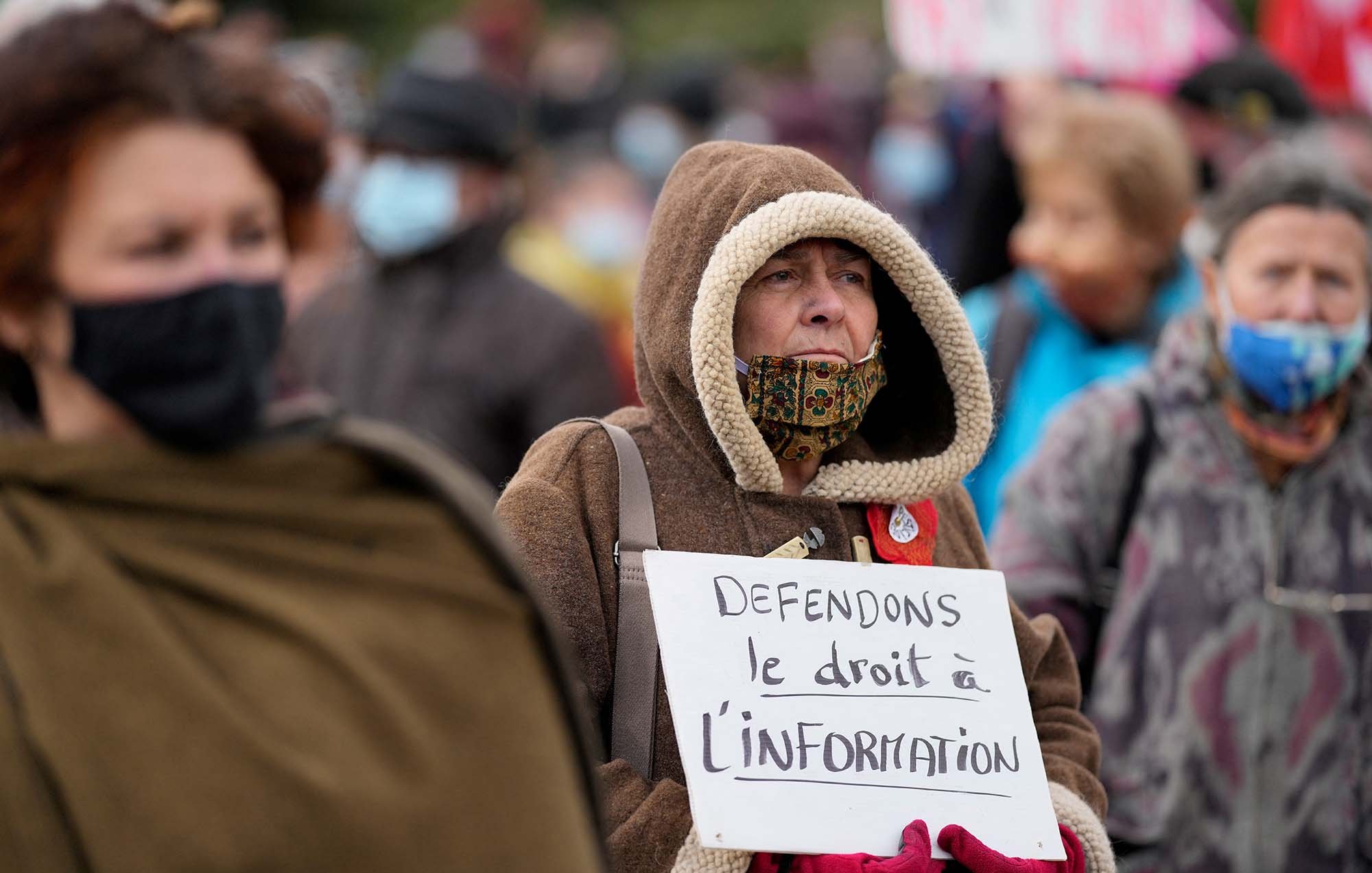L'image montre un groupe de personnes rassemblées lors d'une manifestation. Au premier plan, une femme âgée porte une grande capuche et un masque, tenant une pancarte sur laquelle il est écrit : "Défendons le droit à l'information". Les autres manifestants sont flous en arrière-plan, mais on peut voir qu'ils portent également des masques. L'atmosphère semble sérieuse et engagée, avec des personnes qui militent pour leurs droits.