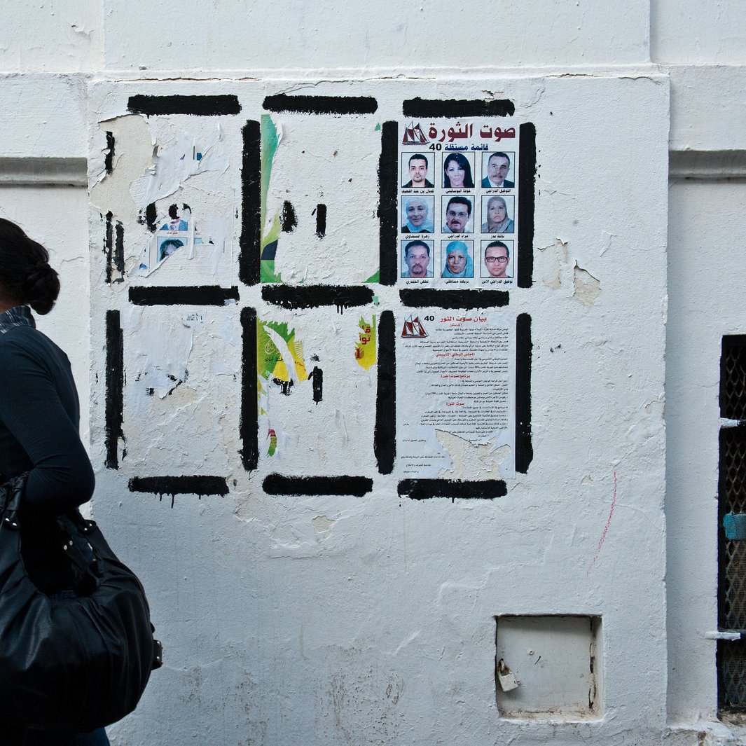 L'image montre une femme marchant le long d'un mur blanc. Elle porte un haut noir et une écharpe. Le mur derrière elle est décoré de divers papiers affichés, y compris des portraits de plusieurs personnes et des textes en arabe. L'atmosphère de la scène semble urbaine et vivante, avec un contraste entre la femme et le mur.