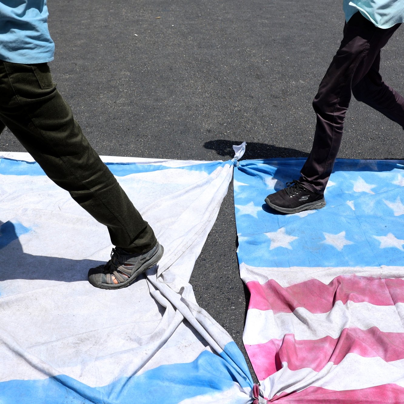 دو نفر در حال قدم زدن روی پرچم‌های دو کشور هستند. زمین زیر آن‌ها خاکی و تاریک است.