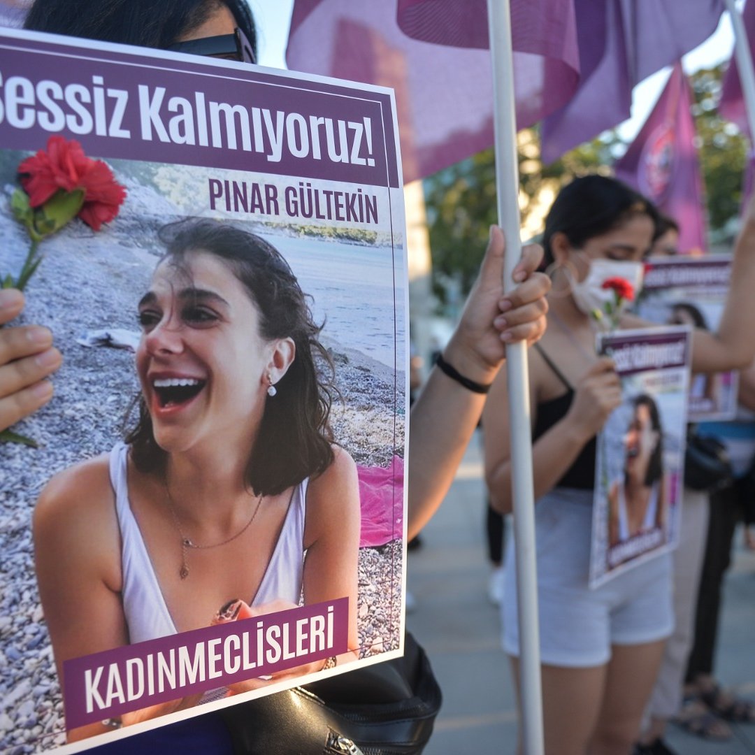L'image montre un groupe de personnes manifestant avec des pancartes. L'une des pancartes porte le visage d'une jeune femme, accompagnée d'un message en turc qui se traduit par "Nous ne restons pas silencieuses !". Une manifestante tient une fleur rouge. Les femmes portent des vêtements variés et semblent engagées dans une cause, probablement liée à la défense des droits des femmes. L'atmosphère peut être perçue comme déterminée et solidaire, avec un accent sur la mémoire d'une personne nommée Pınar Gültekin.