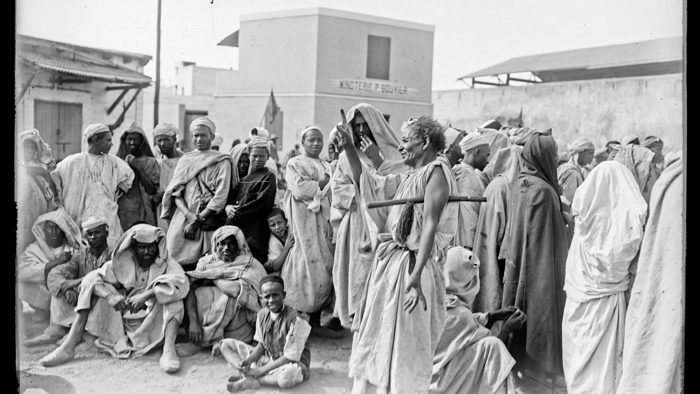 L'image montre un groupe de personnes rassemblées dans un environnement extérieur. Elles portent des vêtements traditionnels et semblent discuter ou observer quelque chose au centre. L'atmosphère est marquée par une certaine sérénité, mais aussi par une curiosité collective. En arrière-plan, on peut distinguer des bâtiments, suggérant un lieu structuré, potentiellement un marché ou un rassemblement communautaire. La lumière naturelle éclaire la scène, ajoutant à l'authenticité de l'ambiance.