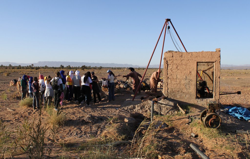 L'image montre un groupe de personnes rassemblées autour d'une structure en terre, possiblement une ancienne construction ou un puits. Il y a une machine ou un mécanisme visible à l'intérieur de la structure. L'environnement est désertique, avec un sol aride et des montagnes en arrière-plan. Le ciel est dégagé, reflétant une ambiance ensoleillée. Les participants semblent s'intéresser à l'activité en cours, observant attentivement.