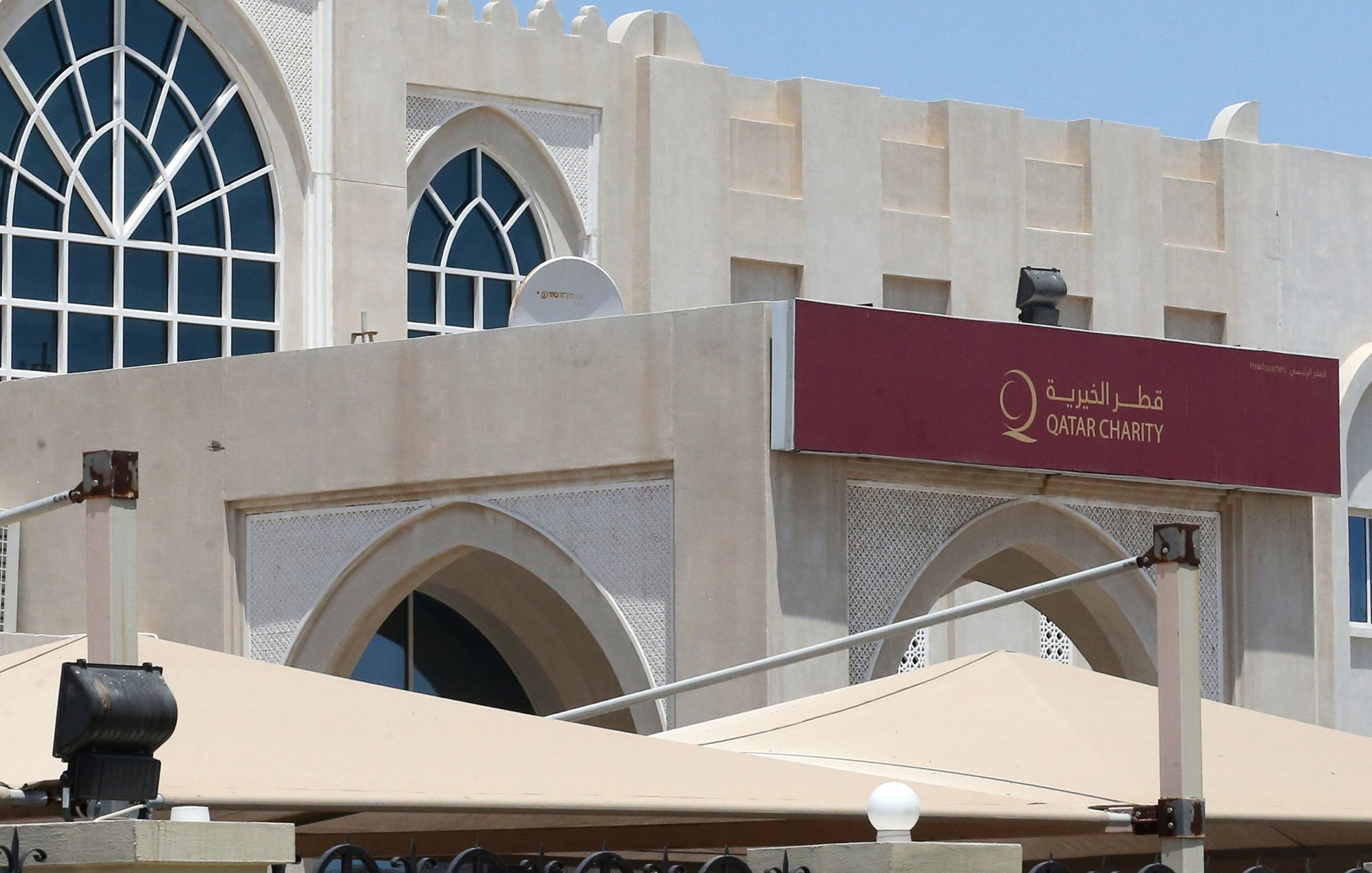L'image montre un bâtiment de style architectural arabe moderne, avec une grande fenêtre en arc sur le mur. Au-dessus de l'entrée, il y a une enseigne en arabe et en anglais avec le nom "Qatar Charity". Le bâtiment est entouré de parasols et d'une clôture, et on peut voir un drapeau du Qatar flottant au sommet du bâtiment. Le ciel est dégagé et ensoleillé.