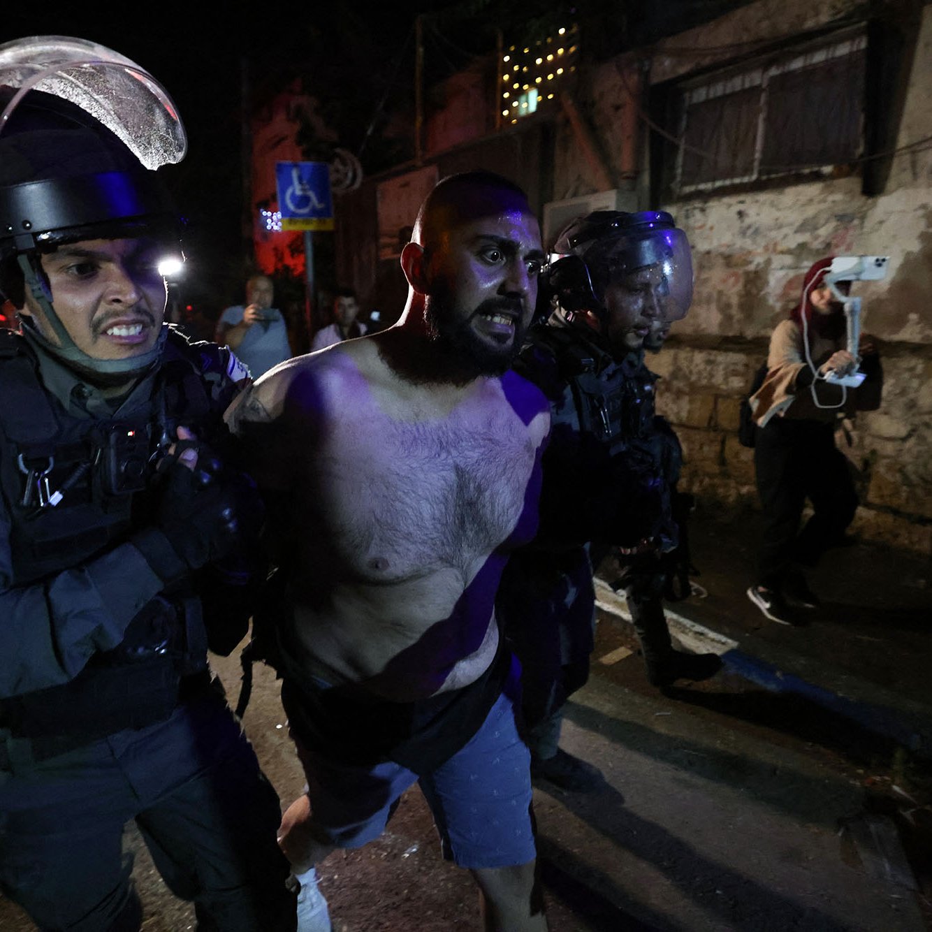 L'image montre des policiers en uniforme, portant des casques et des protections, en train de maîtriser un homme. L'homme est torse nu et semble être en train d'être arrêté. L'environnement nocturne, avec des lumières et des bâtiments en arrière-plan, suggère une situation de tension, potentiellement liée à des manifestations ou des troubles civils. Les expressions et la posture des policiers indiquent une approche ferme envers la situation.